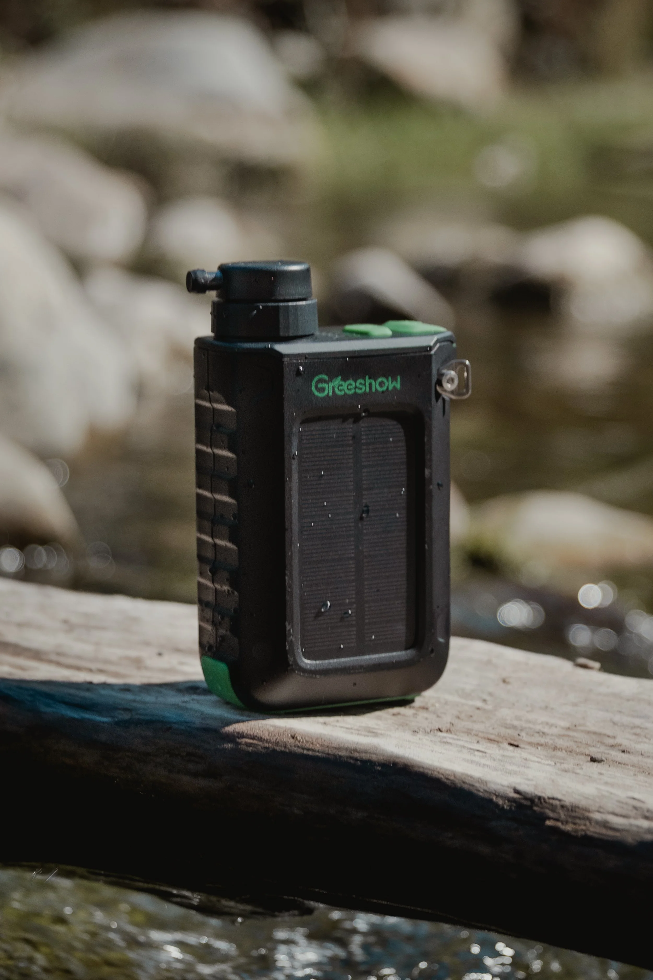 A portable solar-powered device with the brand name 'Greeshow', placed on a wooden log near a body of water, surrounded by rocks.