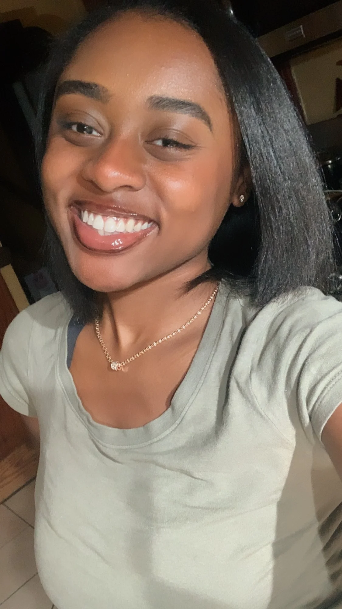 A young woman with black hair, smiling and sticking out her tongue, wearing a beige T-shirt, a gold necklace, and small earrings, in an indoor setting.