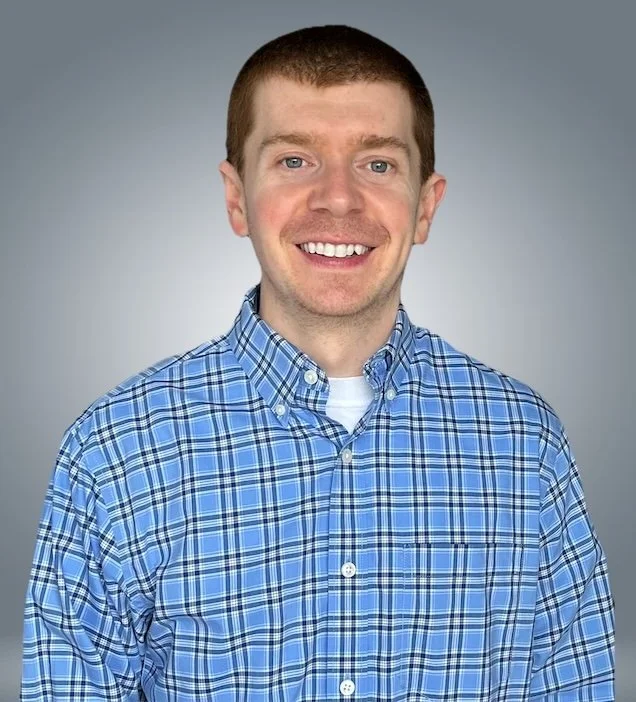 A smiling man with short brown hair wearing a blue plaid button-up shirt and a white T-shirt underneath, against a gray background.