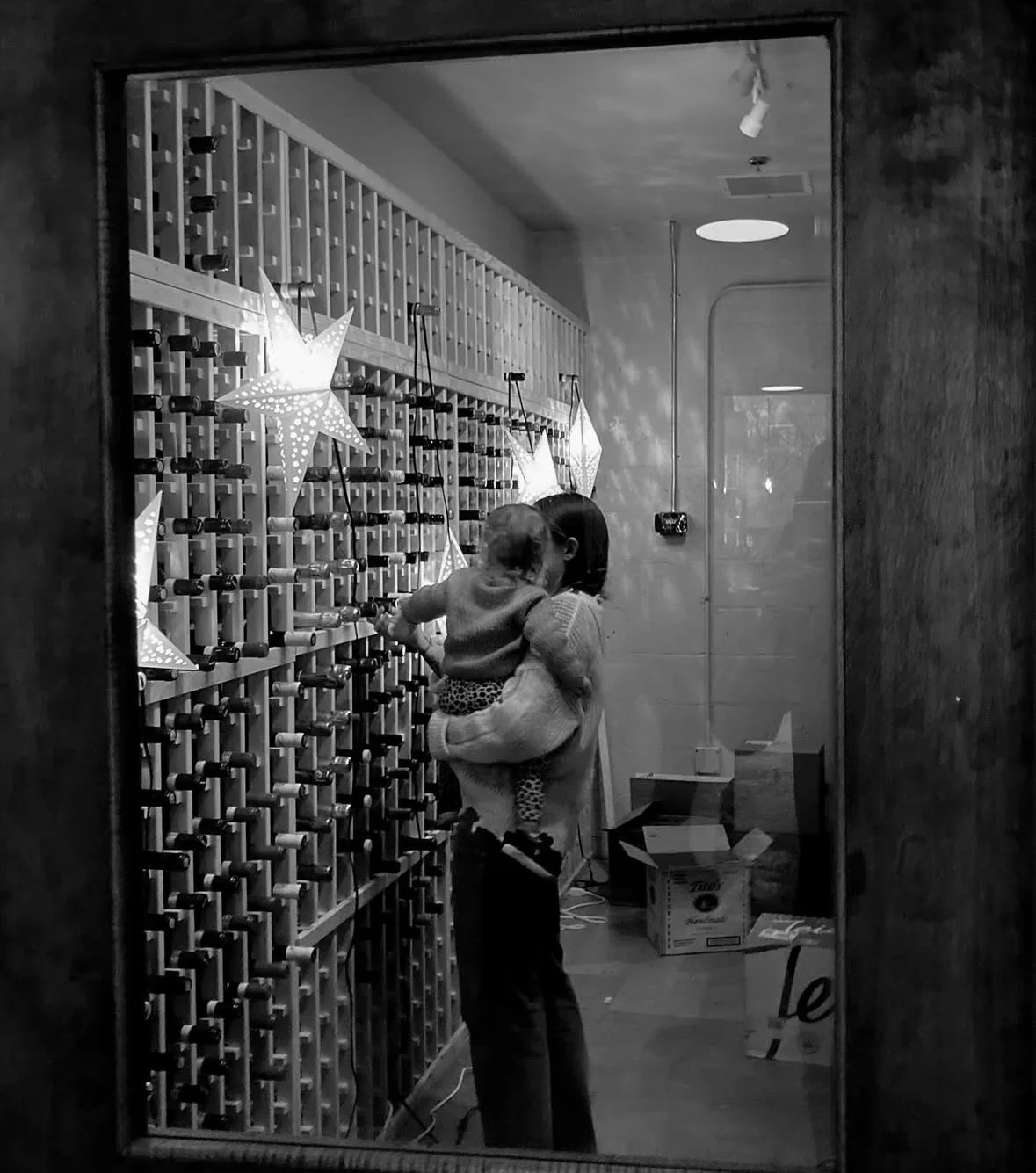 A woman holding a child, standing in front of a wall with star-shaped lights and wine bottle racks, reflected in a mirror.