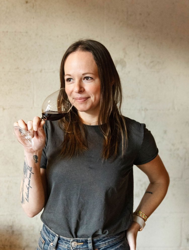 A woman with long brown hair holding a glass of red wine, wearing a dark gray t-shirt and jeans, standing against a light-colored brick wall.