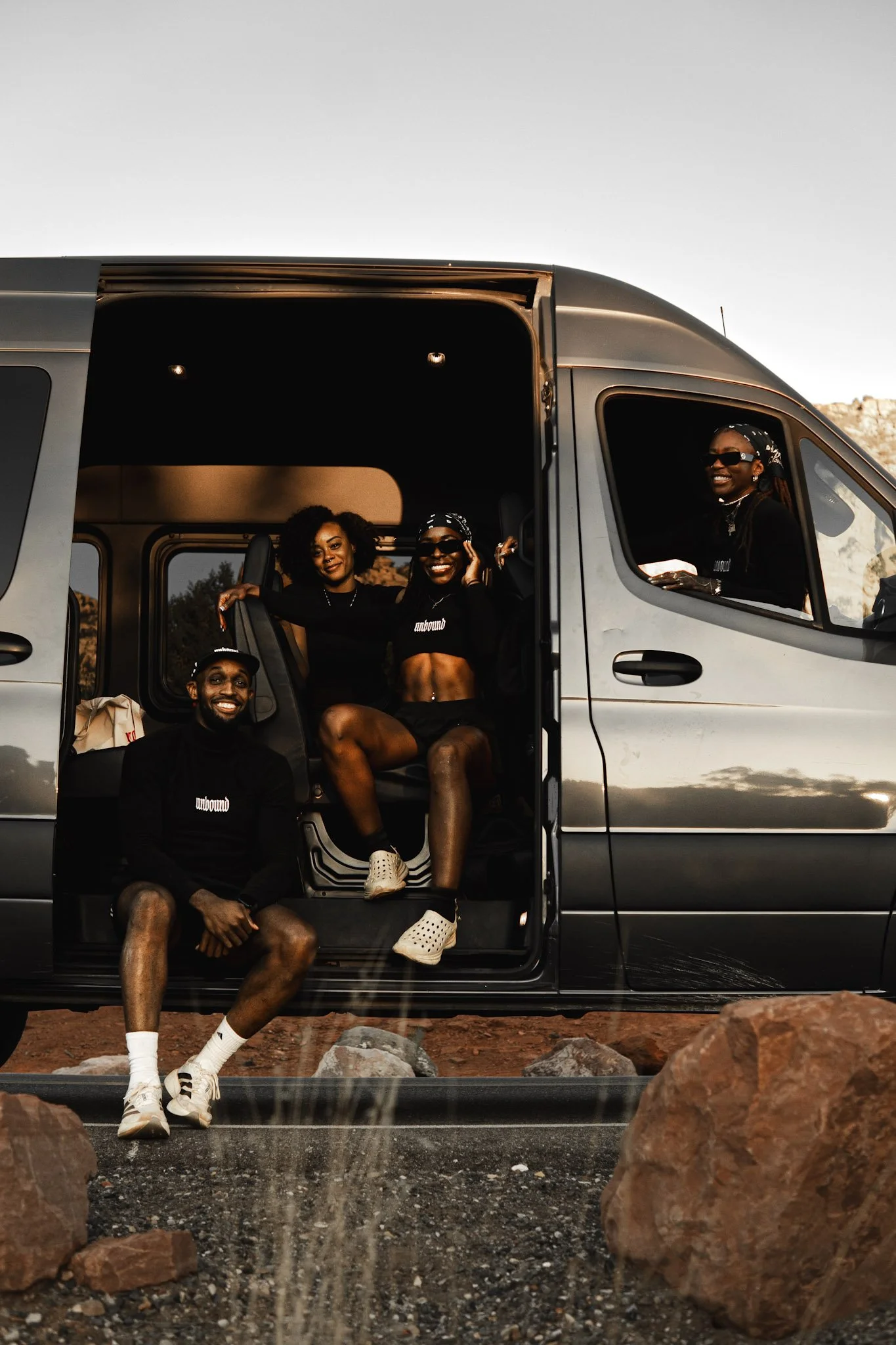 Four people sitting and standing around a van, smiling at the camera outdoors during sunset.