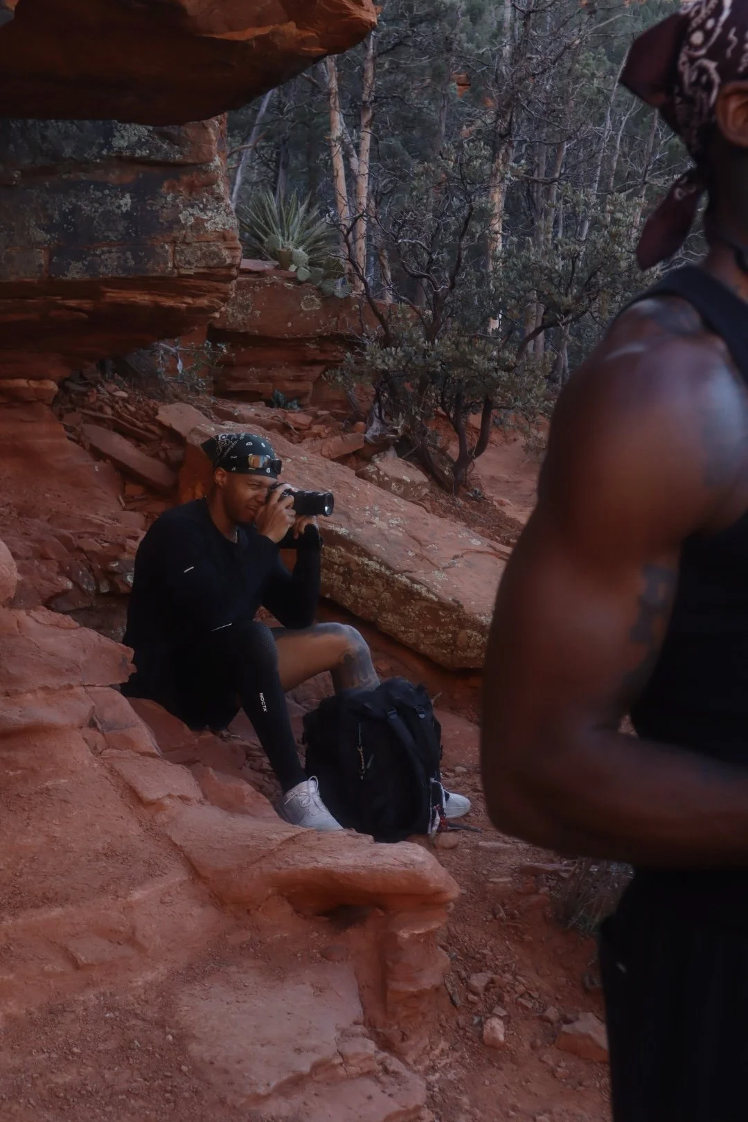 A person sitting on red rocky terrain taking a photo with a camera, wearing a black long-sleeve shirt, black shorts, white sneakers, a black headband, and black knee-high socks. A black backpack is beside them. Part of another person, wearing a black