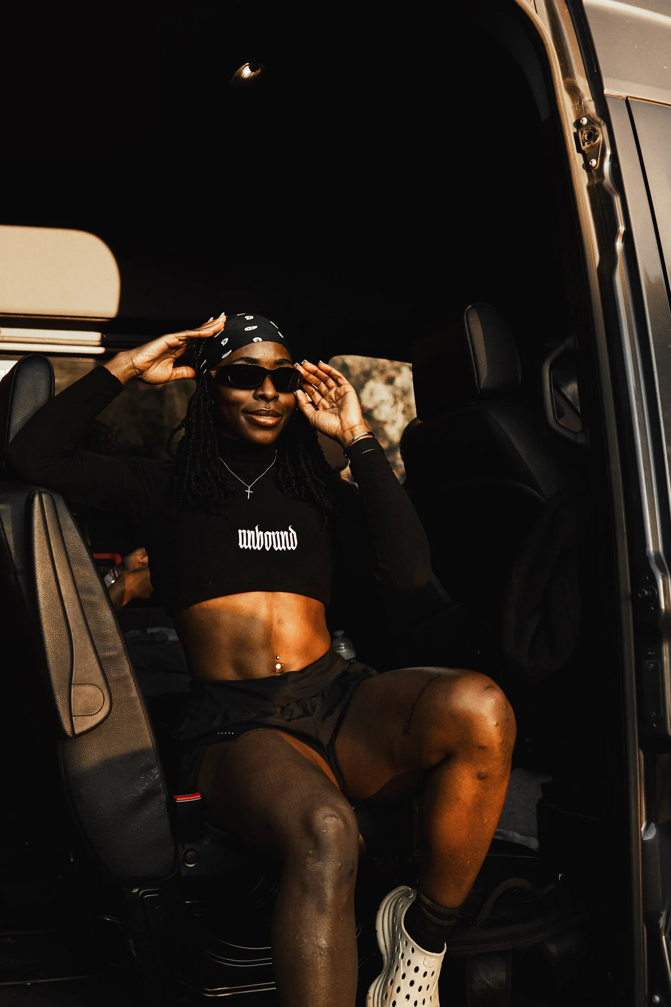 A woman with dark skin sitting in a vehicle, wearing black sunglasses, a black bandana, a black crop top that says 'unbound', black shorts, and white Crocs, adjusting her sunglasses.