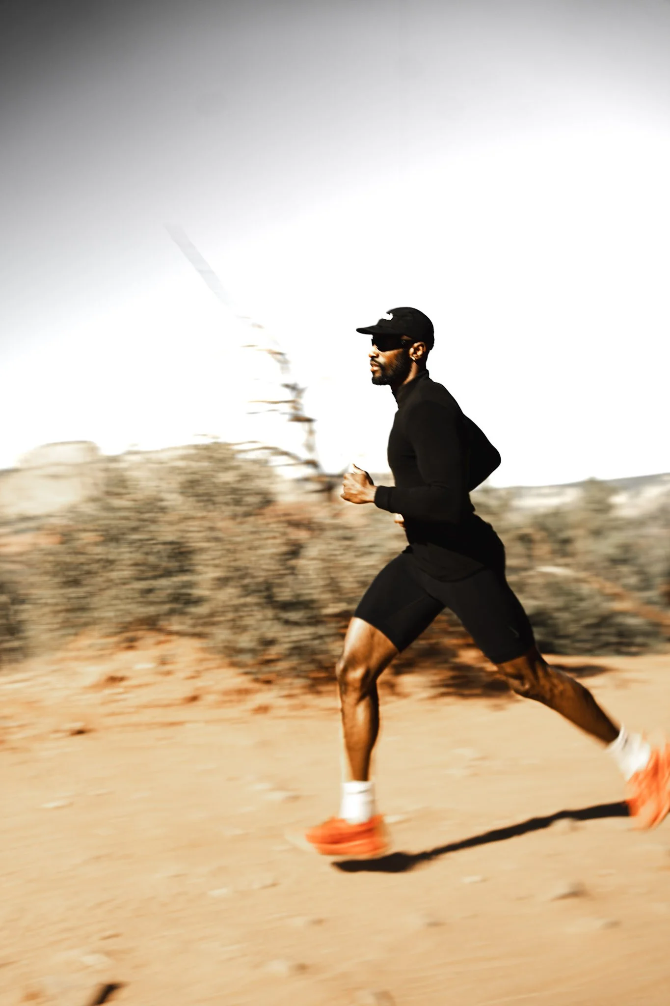 A man running outdoors in a desert landscape, wearing black athletic clothing, a cap, and orange running shoes.