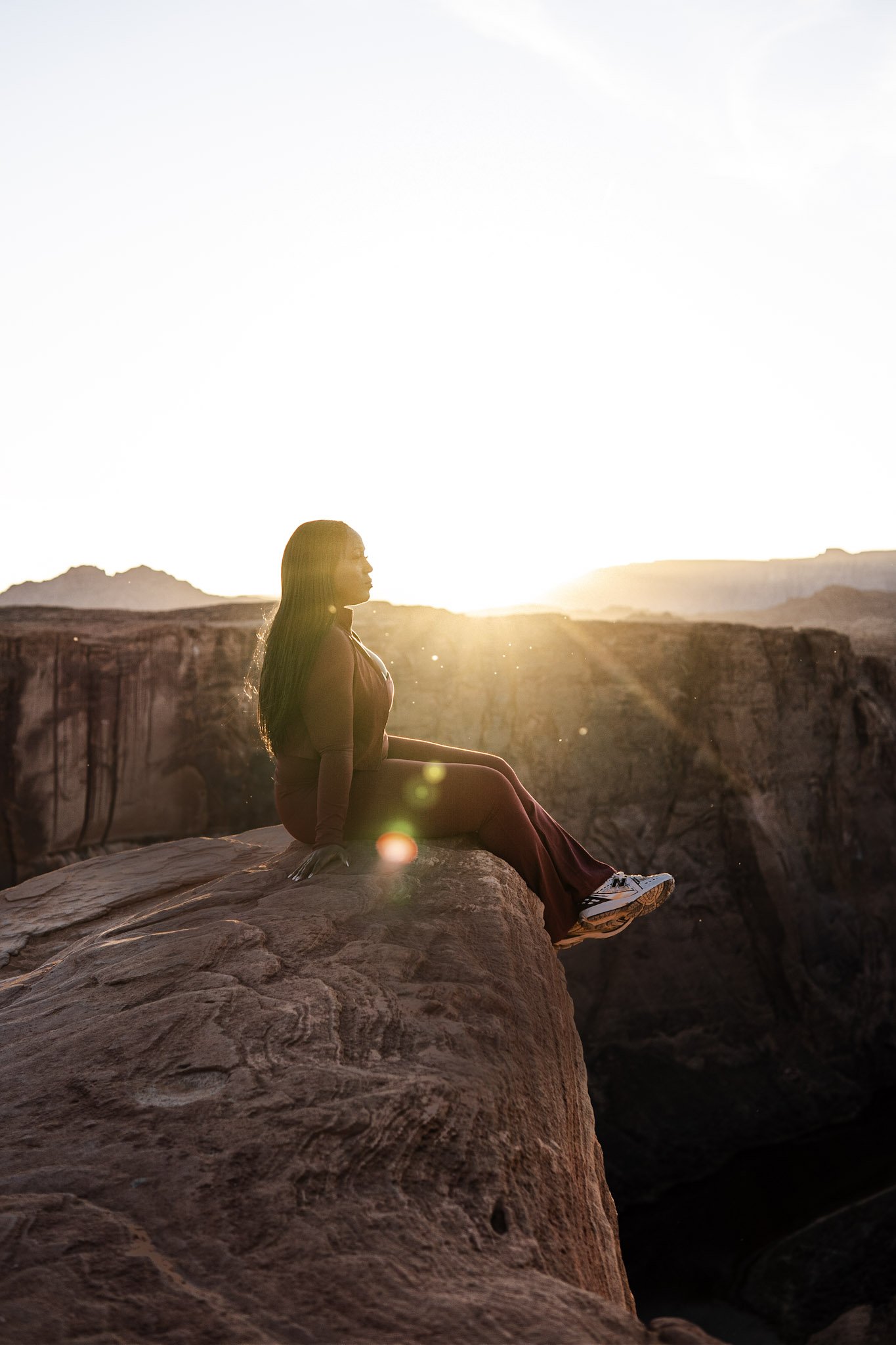 A woman sitting on the edge of a rocky cliff during sunset, with the sun behind her and a landscape of distant mountains in the background.