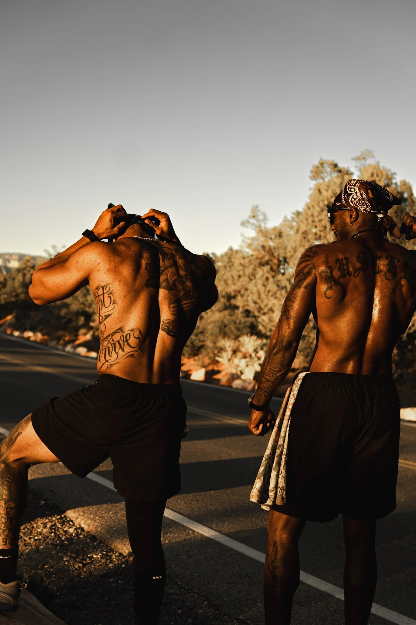 Two shirtless men with tattoos walking on a road during sunset, with trees in the background. One man is adjusting his headwear, while the other is wearing a bandana and glasses.