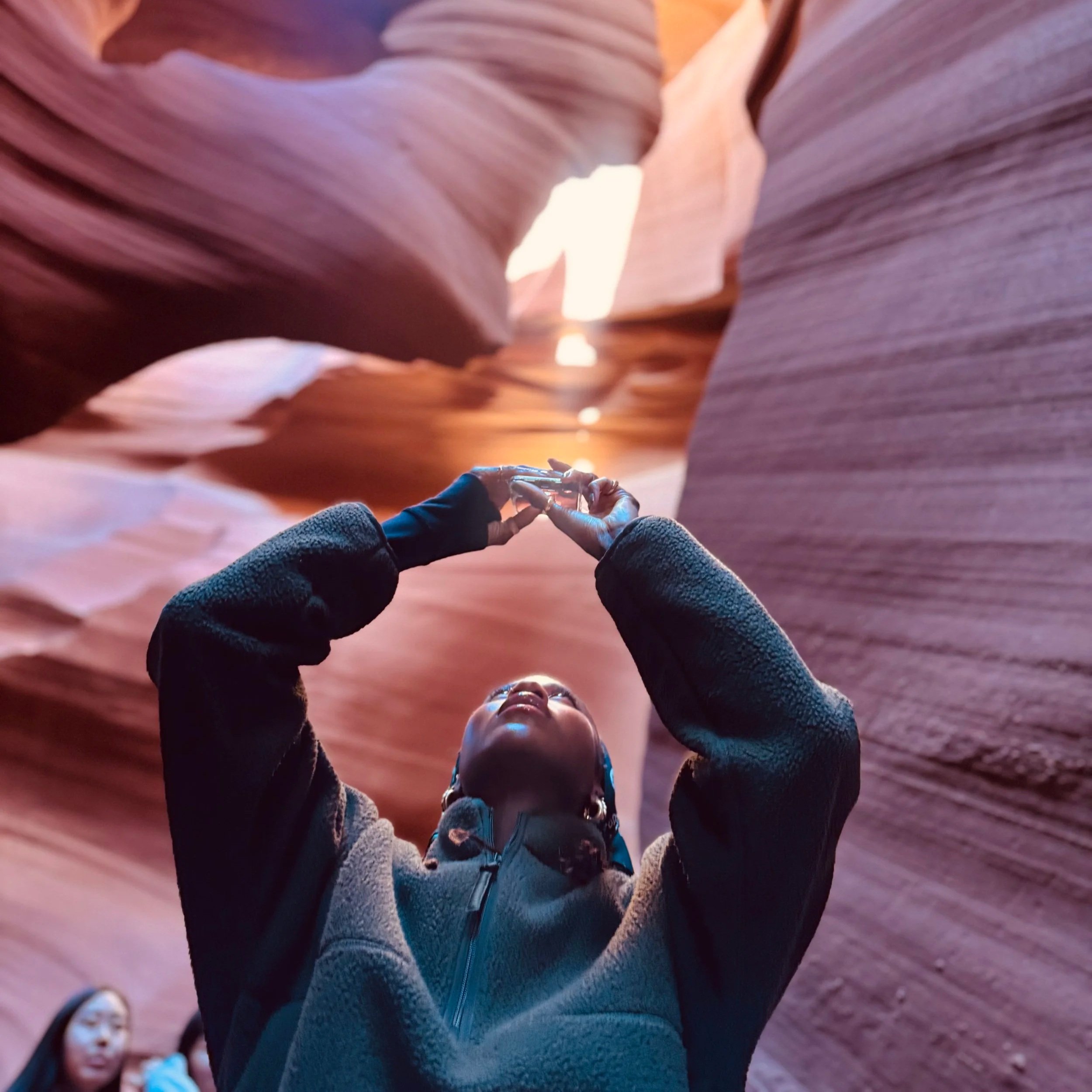 A person with a dark hoodie and head covering looks up while gesturing with hands in a narrow canyon with smooth, layered sandstone walls.