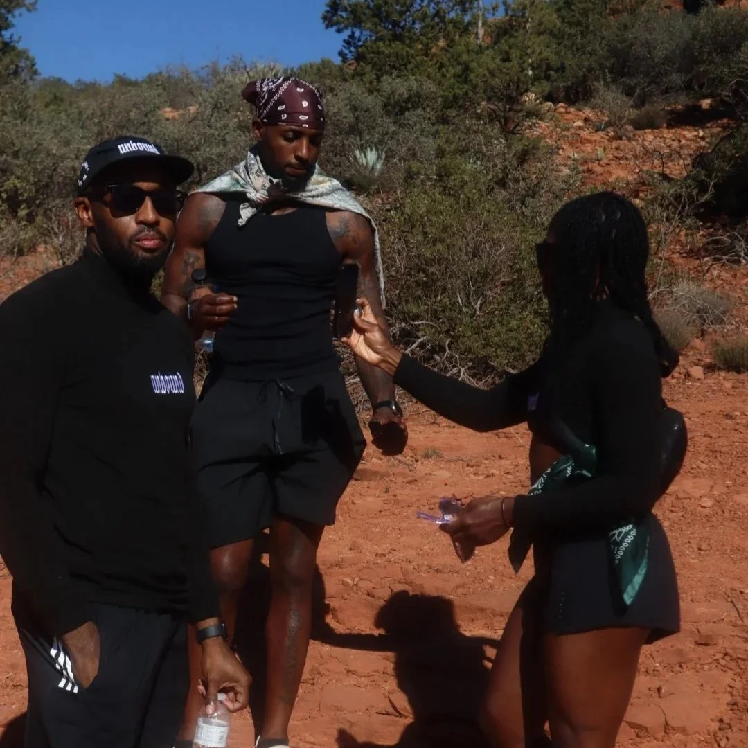 Three people standing outdoors on a dirt trail with shrubs and trees in the background. The person in the middle is taller, wearing a black tank top, black shorts, and a patterned bandana. The person on the left is wearing sunglasses, a black cap, an