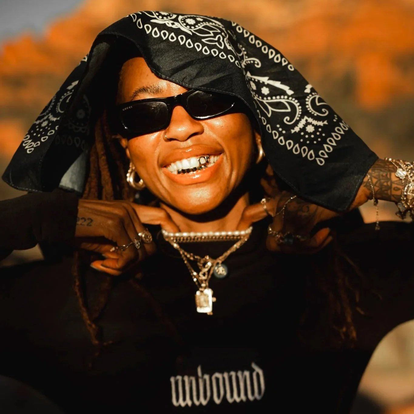 A smiling woman wearing sunglasses, a black bandana with white patterns, and layered jewelry including necklaces, earrings, and bracelets. She is in a close-up outdoor shot with warm sunlight in the background.