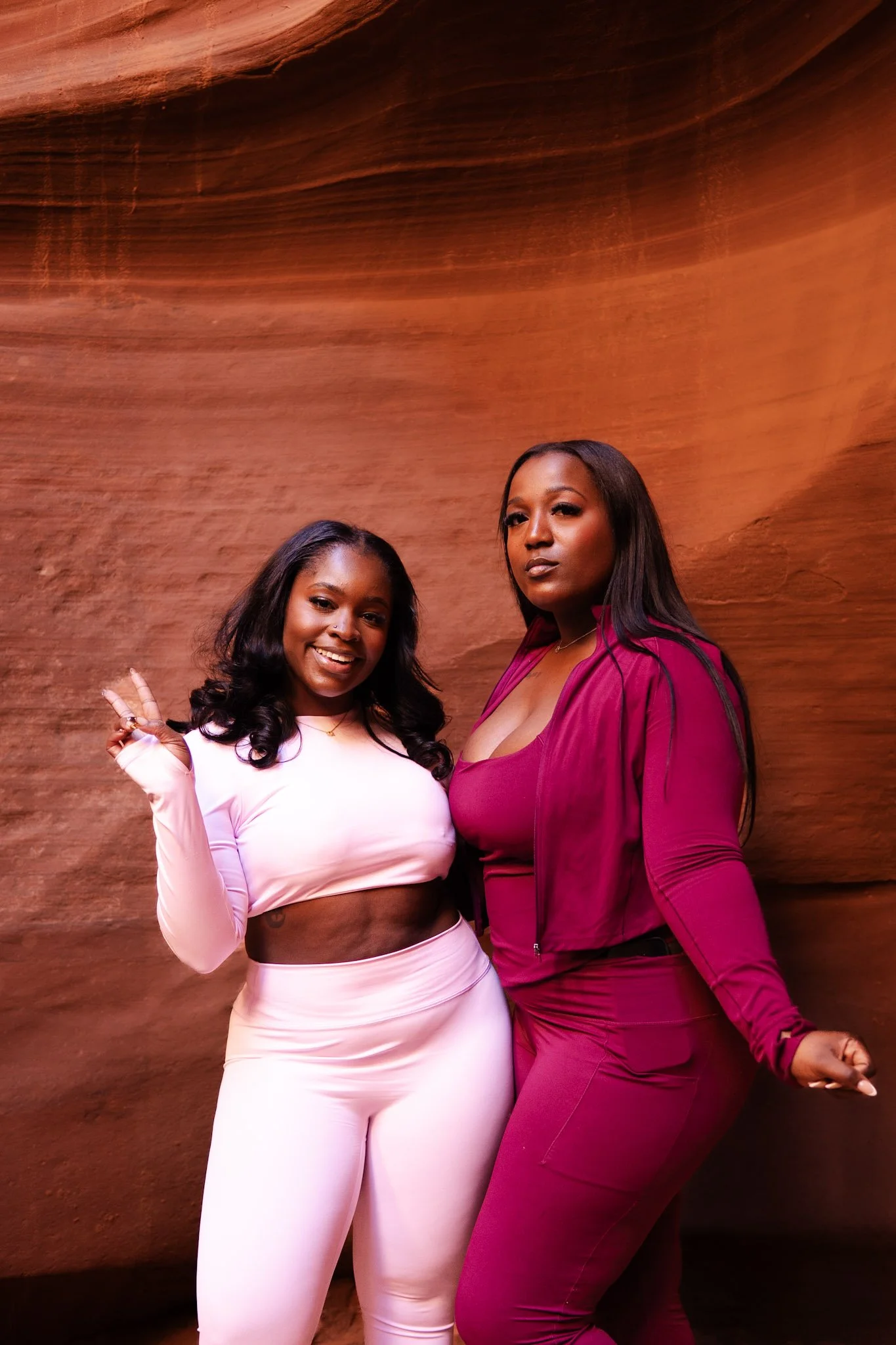 Two women posing in front of a red rock canyon wall, one smiling and making a peace sign, the other with a serious expression, both dressed in pink and maroon athletic outfits.