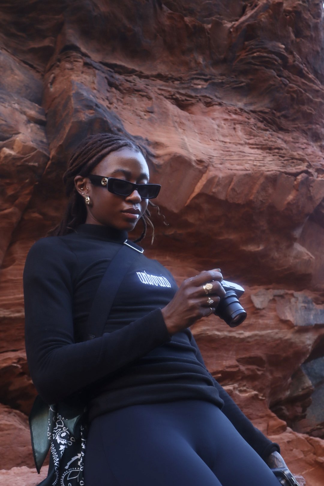 A woman with dreadlocks wearing black sunglasses, gold earrings, and a black long-sleeve shirt with white text, standing in front of red rock formations while holding a camera.