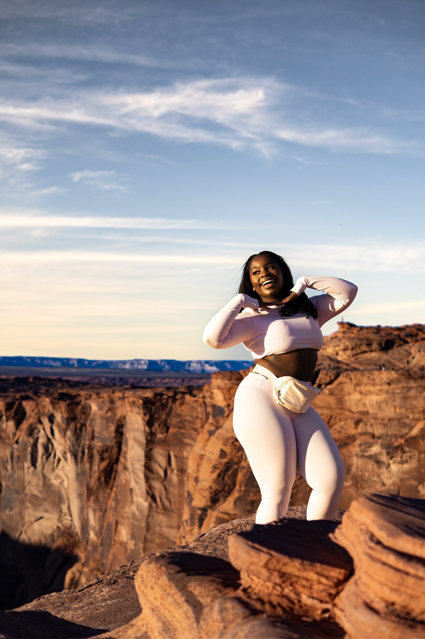 A smiling woman in white athletic clothing standing on a rocky ledge in a canyon during sunset.