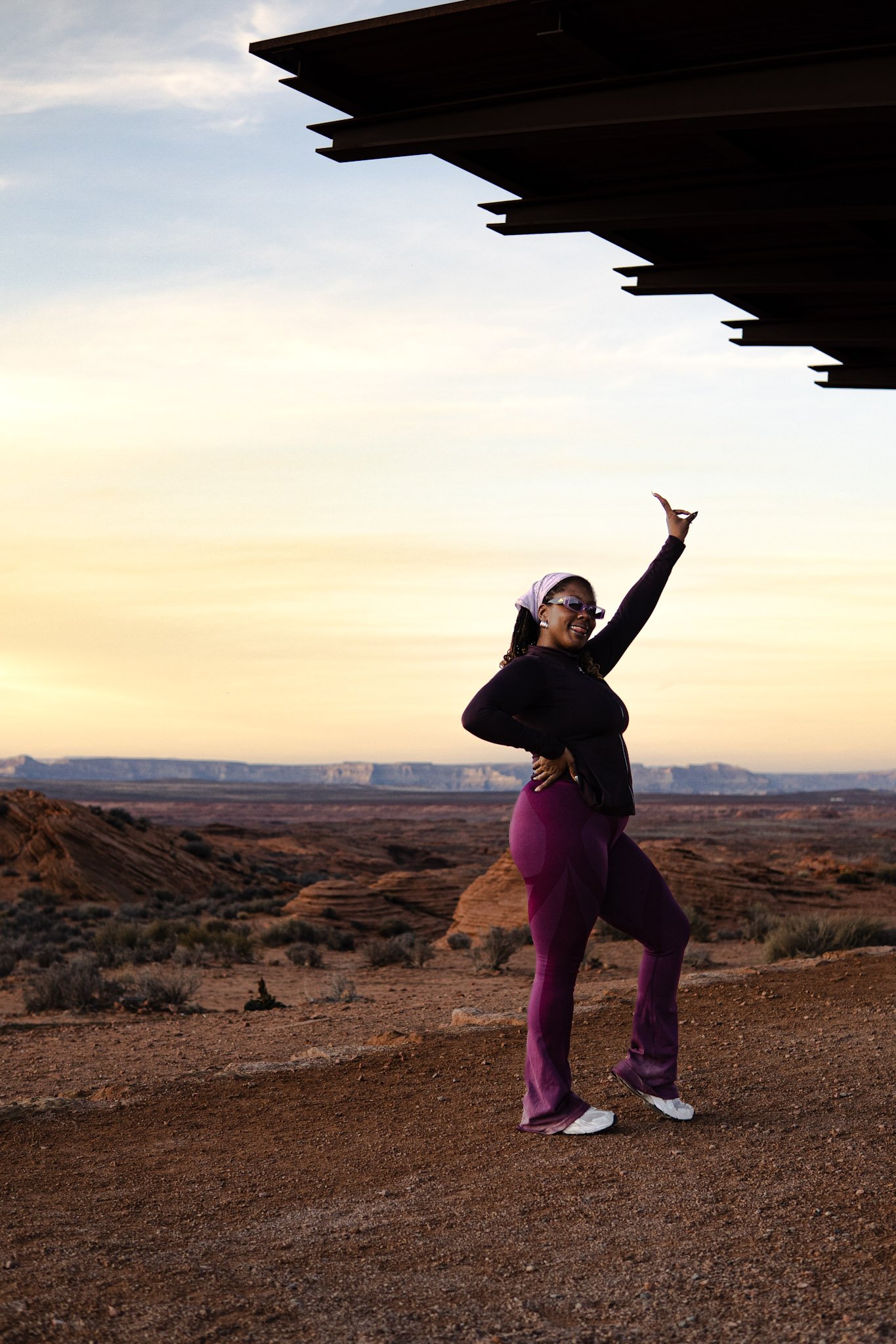 A woman standing in a desert landscape at sunset, striking a pose with one arm raised and the other on her hip, wearing sunglasses, a headscarf, black top, and purple leggings, with a modern building overhang above her.