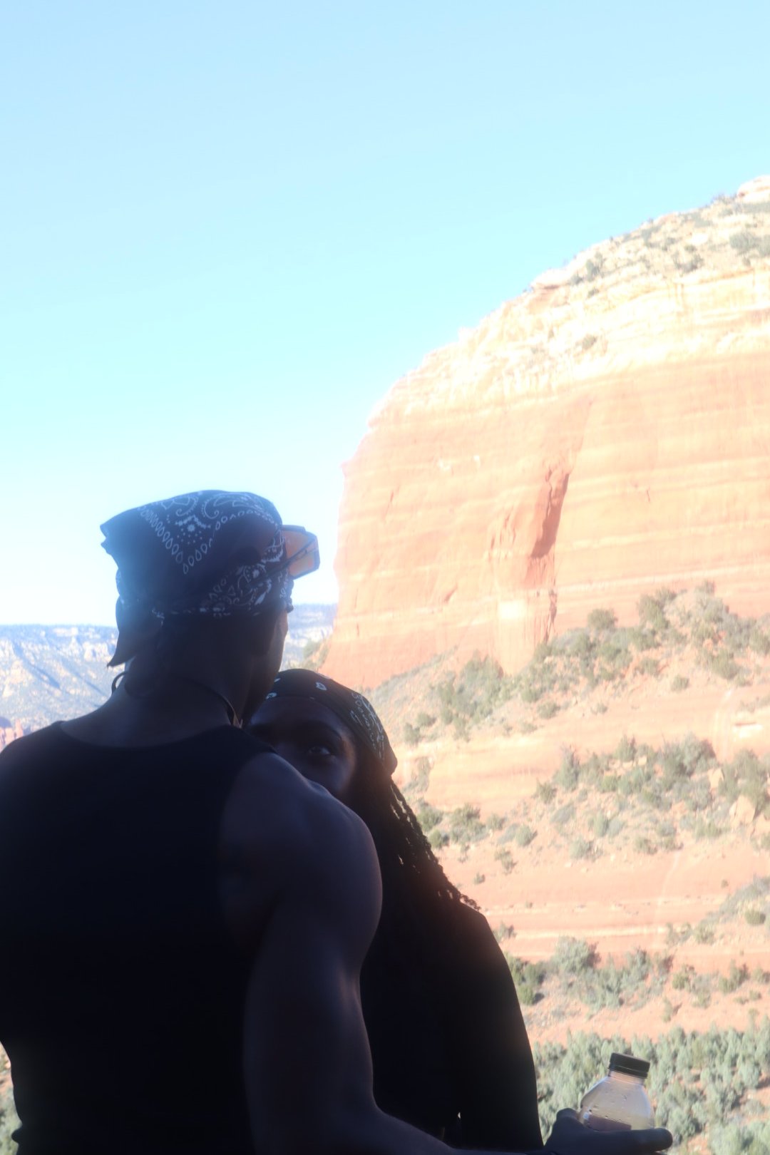 Two people with bandanas on their heads, one holding a water bottle, are outdoors with red rock formations and a clear blue sky in the background.