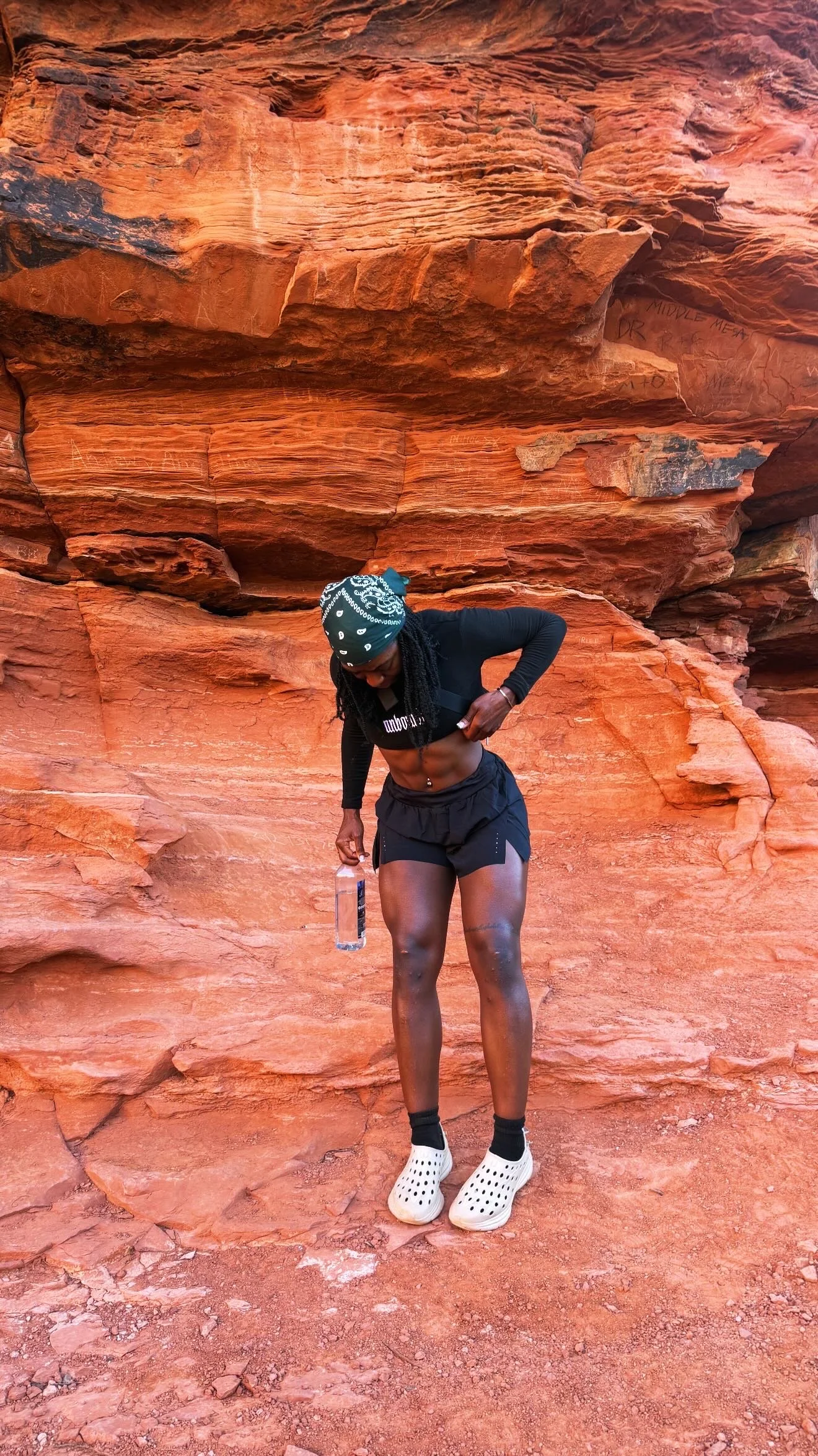 A woman in black athletic clothing, with a bandana, standing in front of red rock formations, holding a water bottle, and lifting her shirt to reveal her toned abdomen.