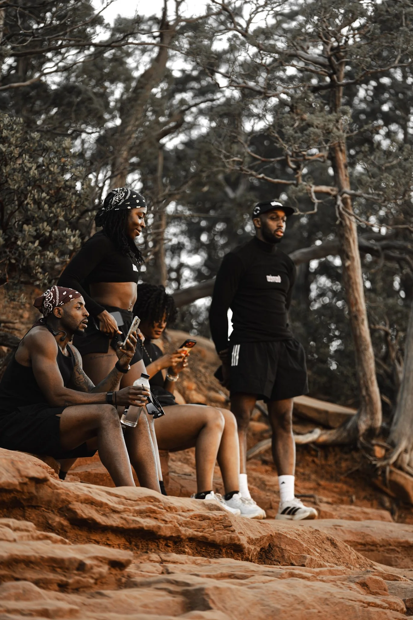 Four young adults in athletic clothing and bandanas, sitting and standing on rocky terrain surrounded by trees, with three of them looking at their phones.