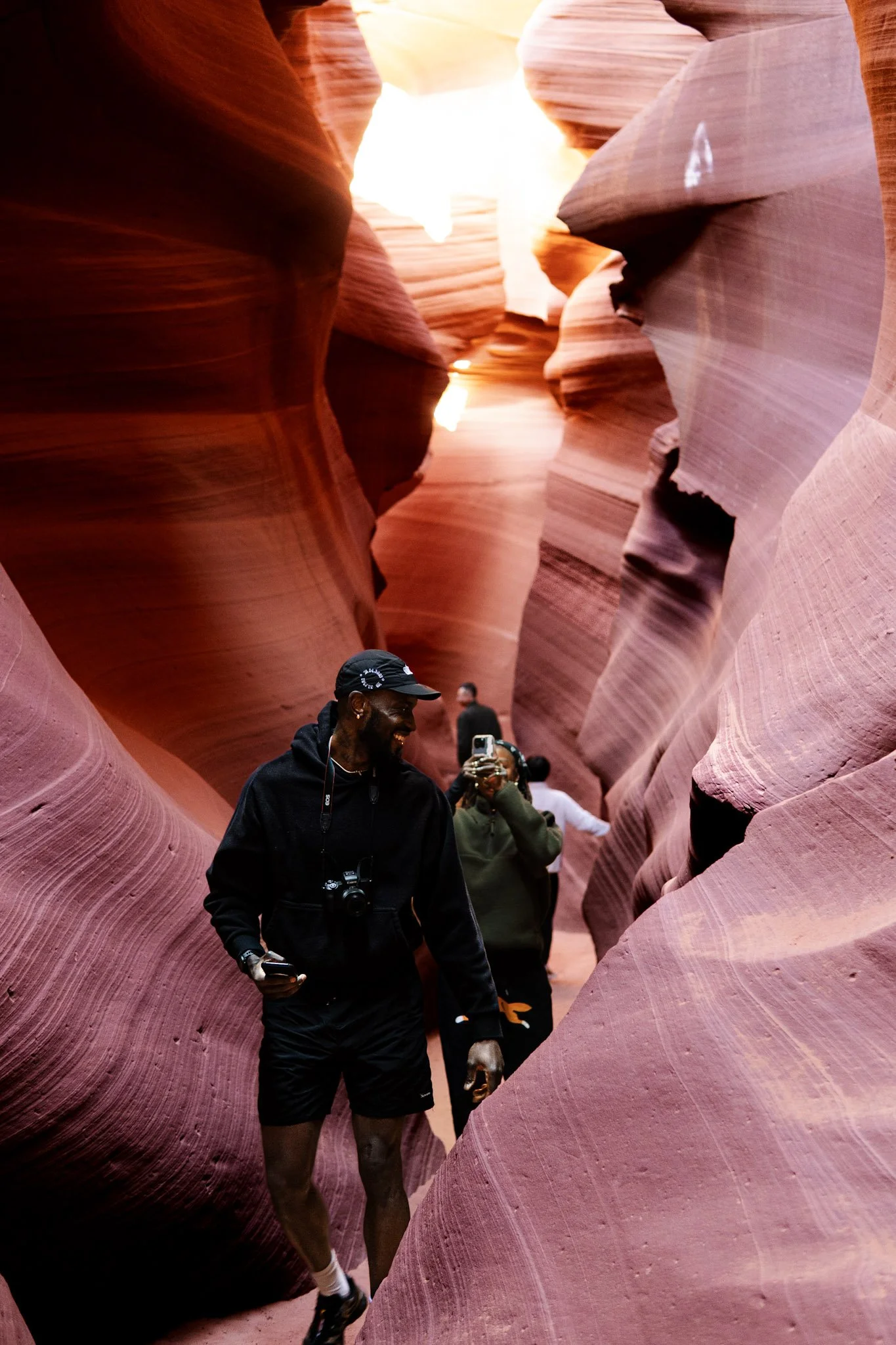 People exploring a narrow slot canyon with curved, reddish sandstone walls, some taking photos, during daylight.