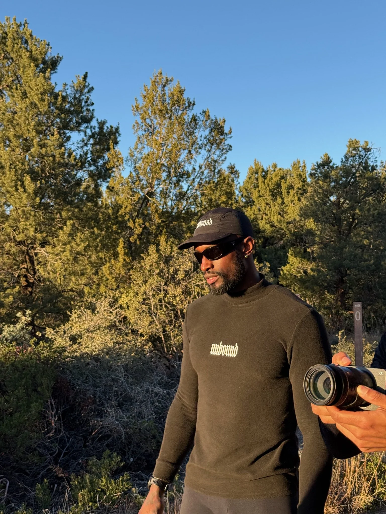 A man wearing sunglasses, a black cap, and a long-sleeve green shirt stands outdoors among trees, holding a camera in his hand.