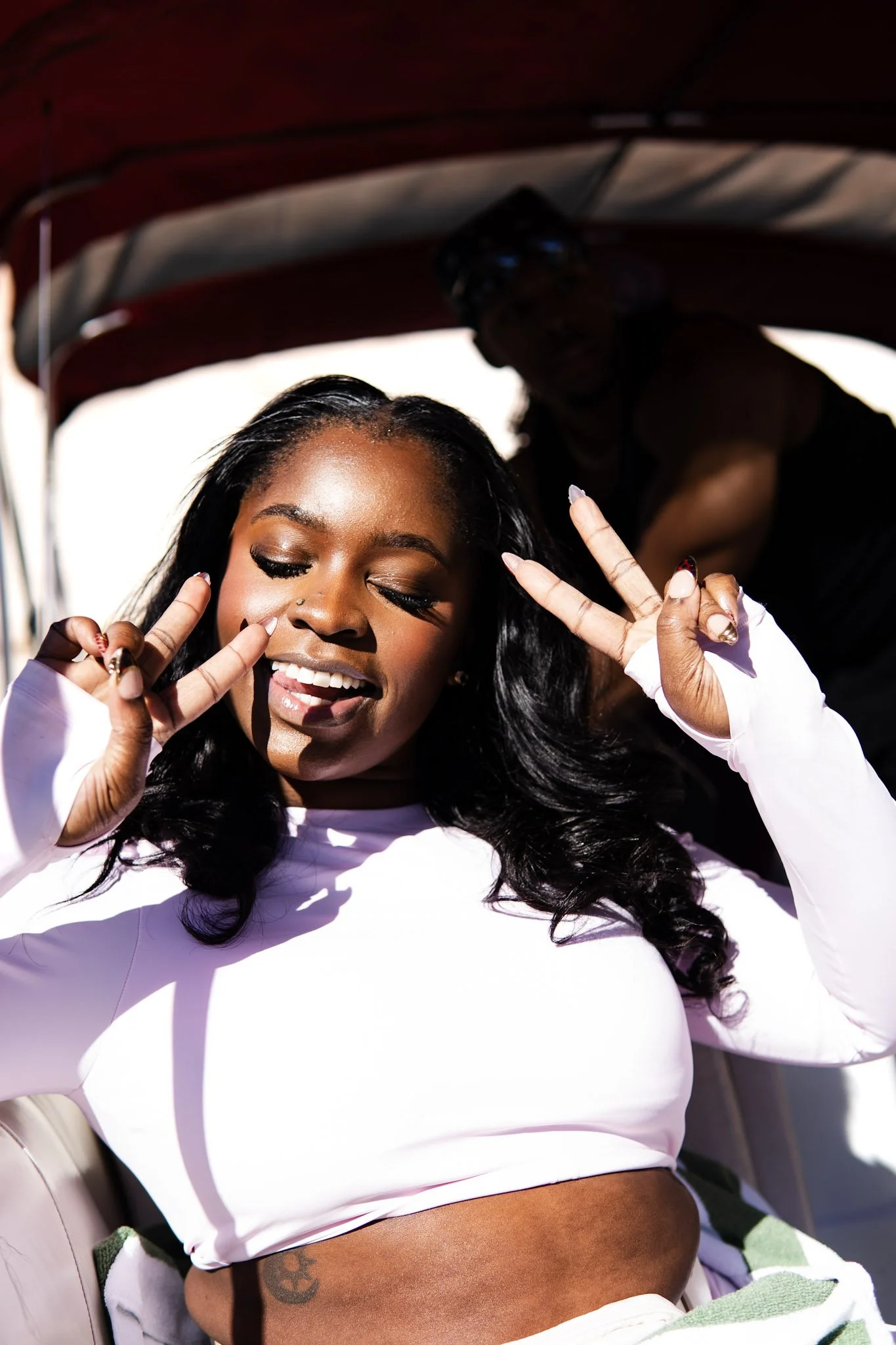 A woman with long black hair and a nose piercing making peace signs with both hands, smiling with her eyes closed, wearing a white long-sleeve top, sitting in a vehicle with an outdoor background. A person is blurred in the background.