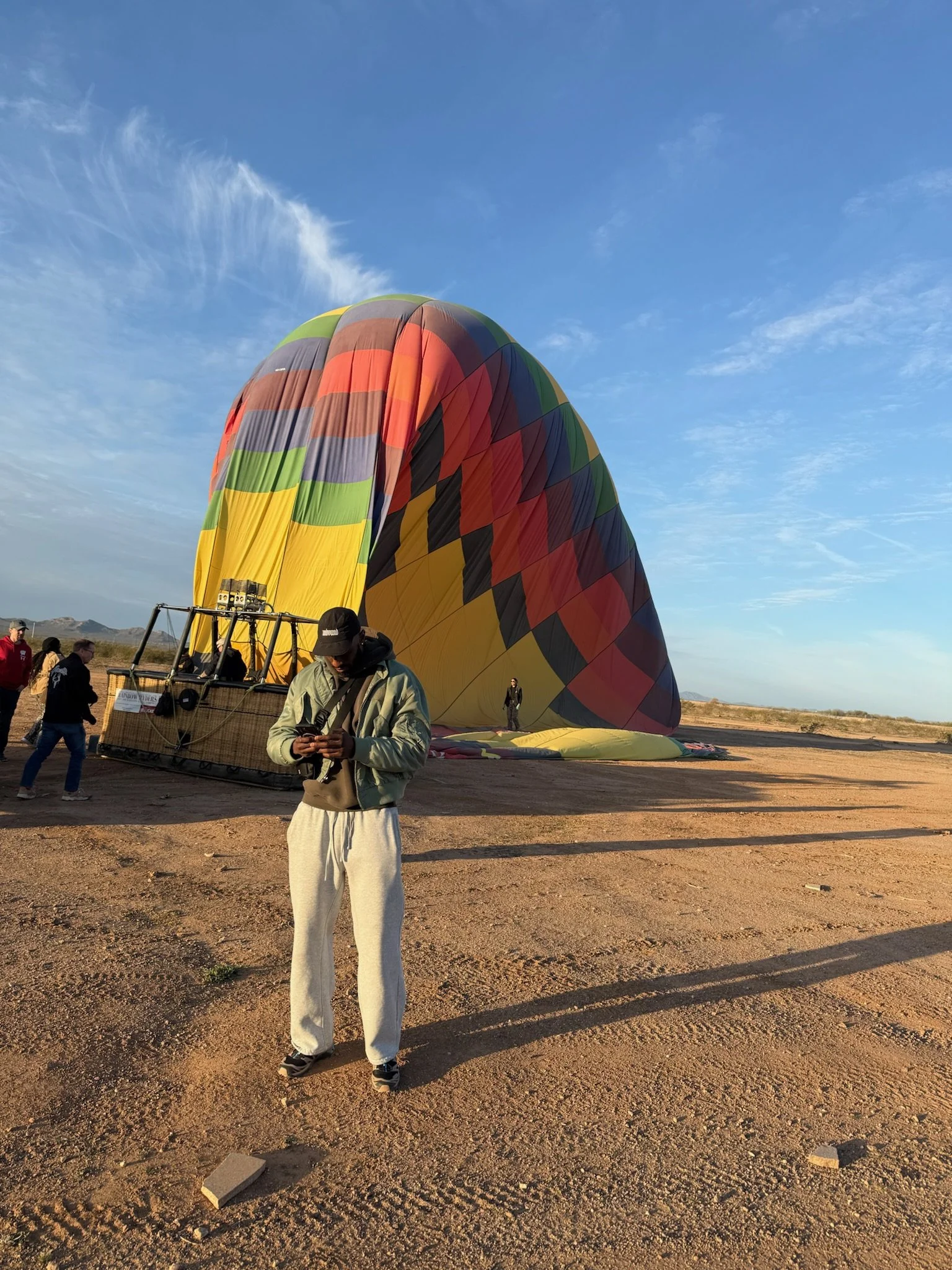 A person in a hoodie and sweatpants looking at a mobile phone in front of a colorful hot air balloon on the ground, with more people in the background and open land under a blue sky.