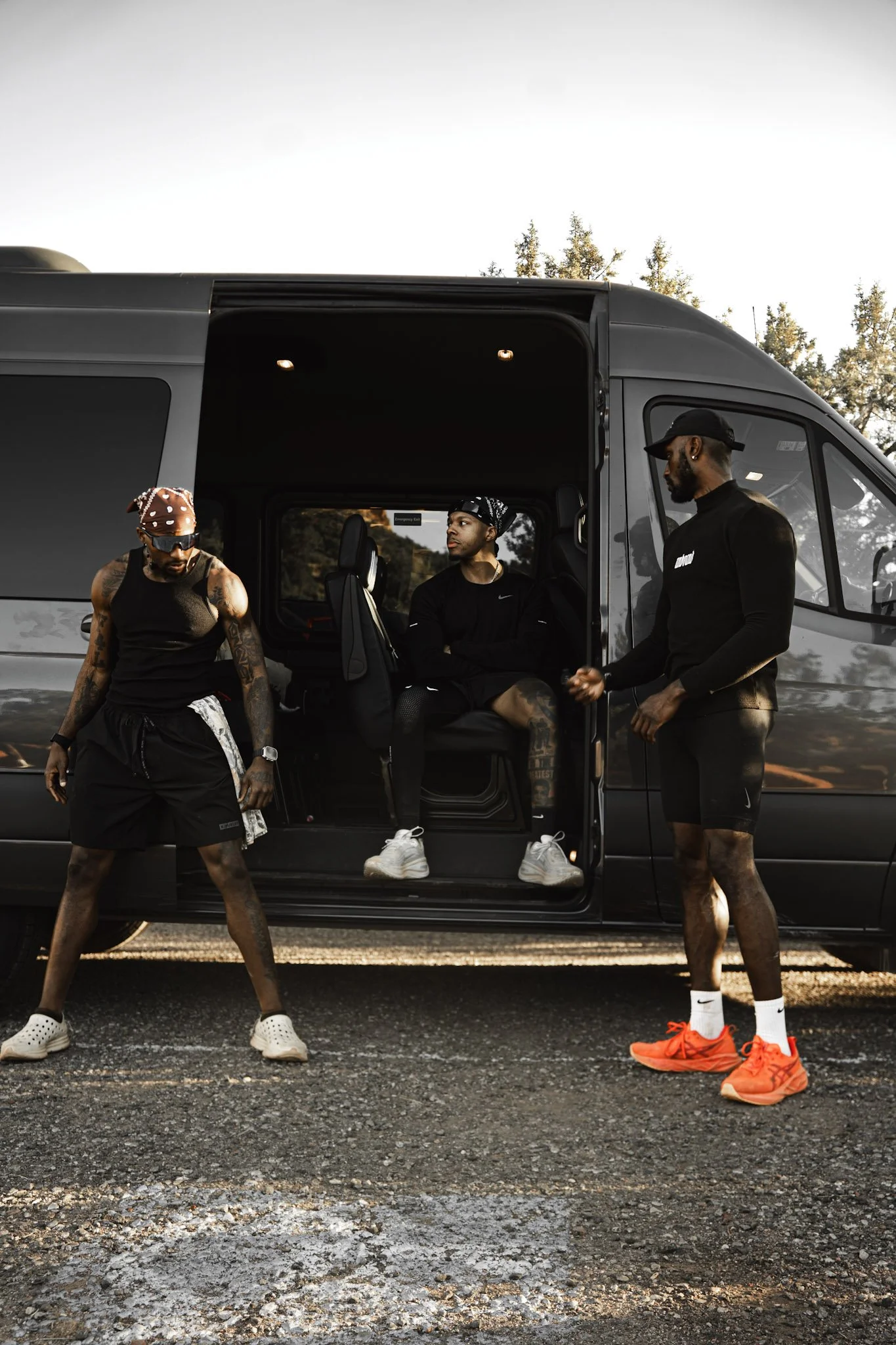 Three men wearing black athletic clothing and sneakers are near a black van parked outdoors. One man is sitting inside the van, while the other two are standing outside, engaged in conversation.