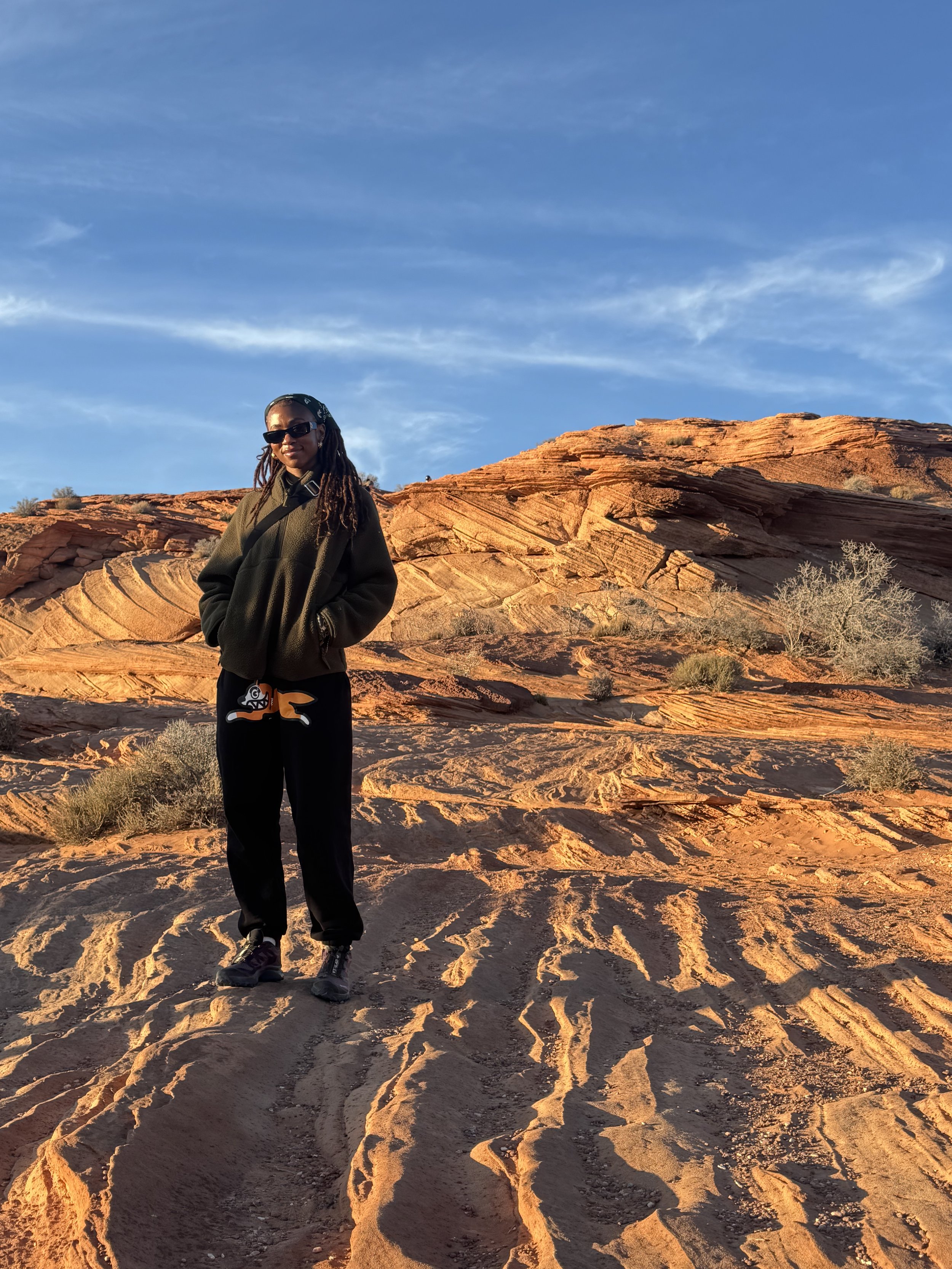 Woman standing on red sandstone desert landscape with blue sky and wispy clouds in the background, wearing sunglasses, a dark jacket, and pants.