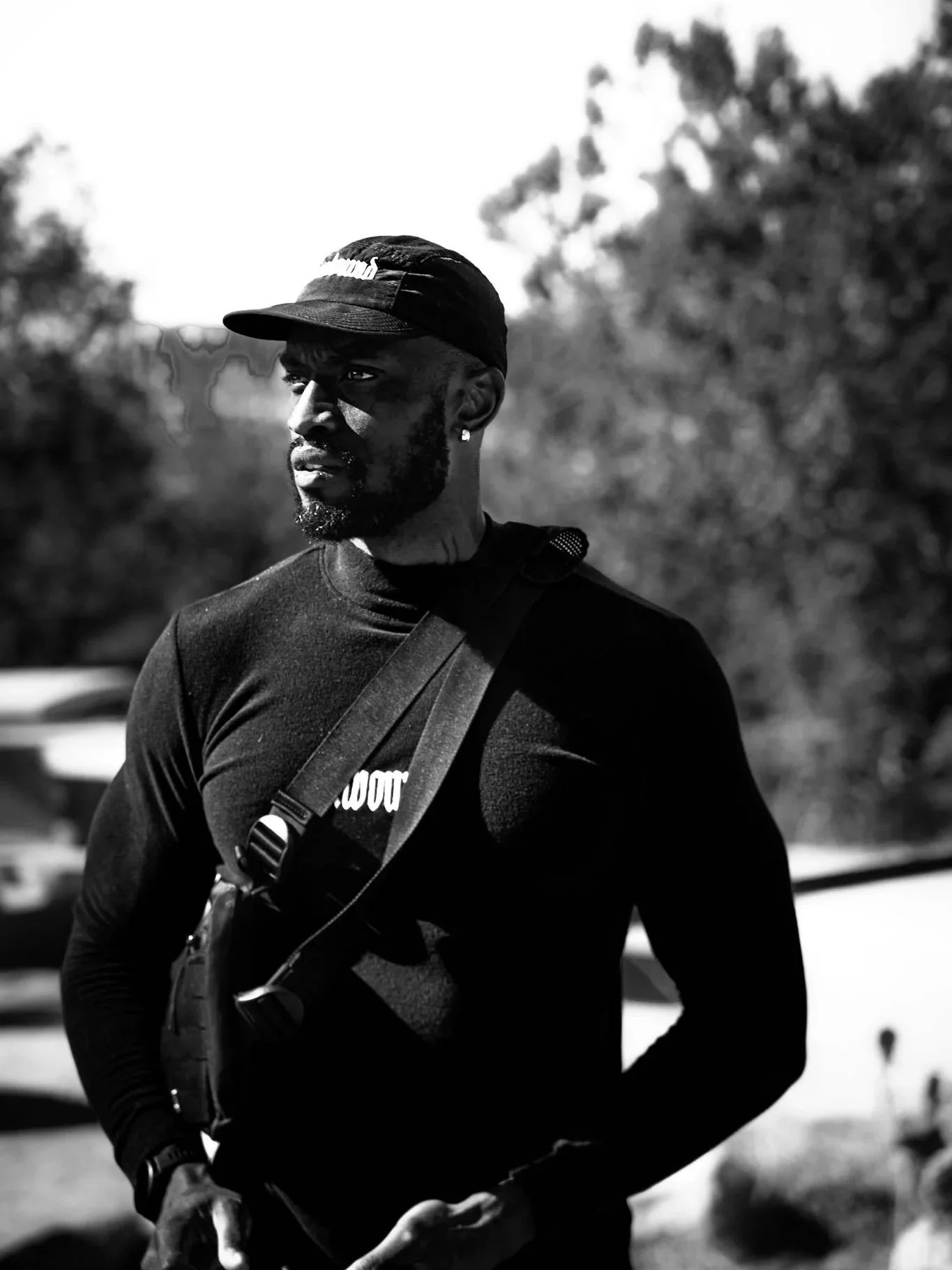 A Black man dressed in athletic clothing, wearing a cap and carrying a camera strap, standing outdoors with trees in the background.