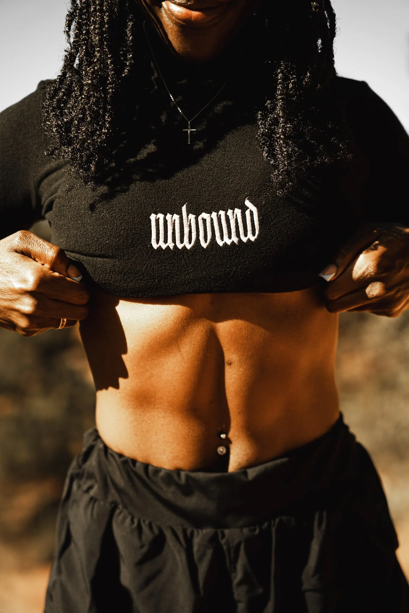 Person lifting black shirt to reveal toned abdomen and piercings, with sunlight highlighting the abdomen, outdoors.