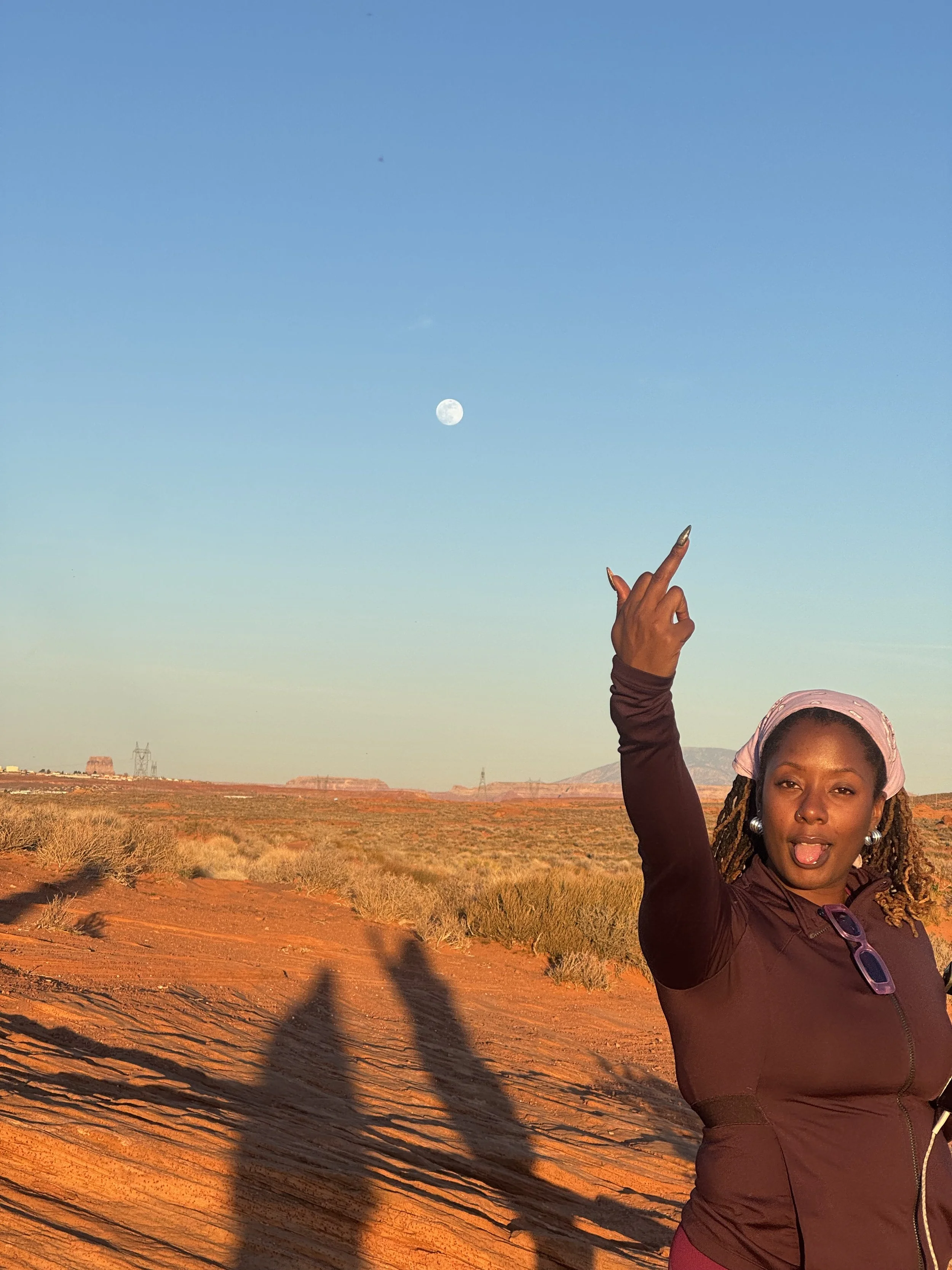 A woman with dreadlocks, wearing a pink headscarf and sunglasses, standing in a desert landscape during sunset, pointing upwards at the moon in a clear sky.
