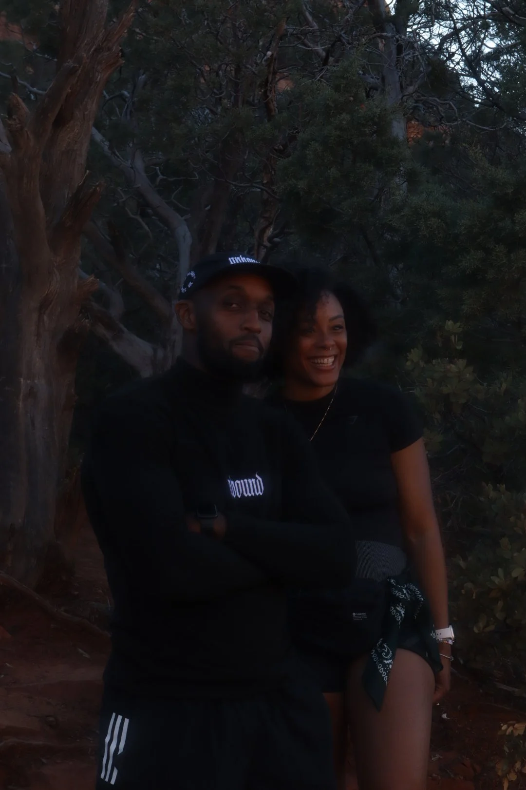 A man and woman posing together outdoors near trees, with the man crossing his arms and the woman laughing.