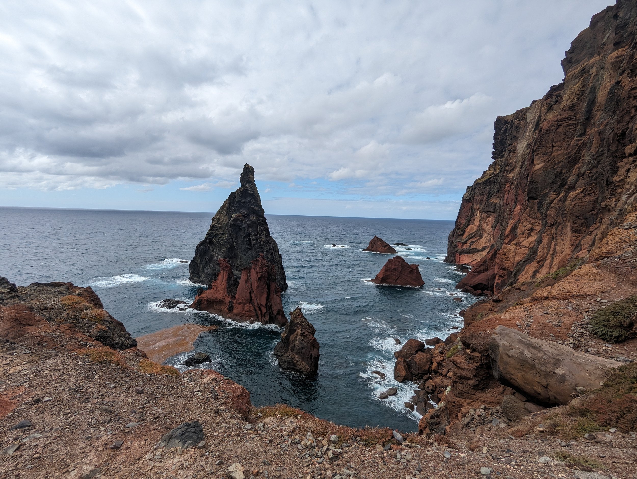 Rocky coastline with large reddish-brown cliffs and several jagged sea stacks in the ocean under a cloudy sky.