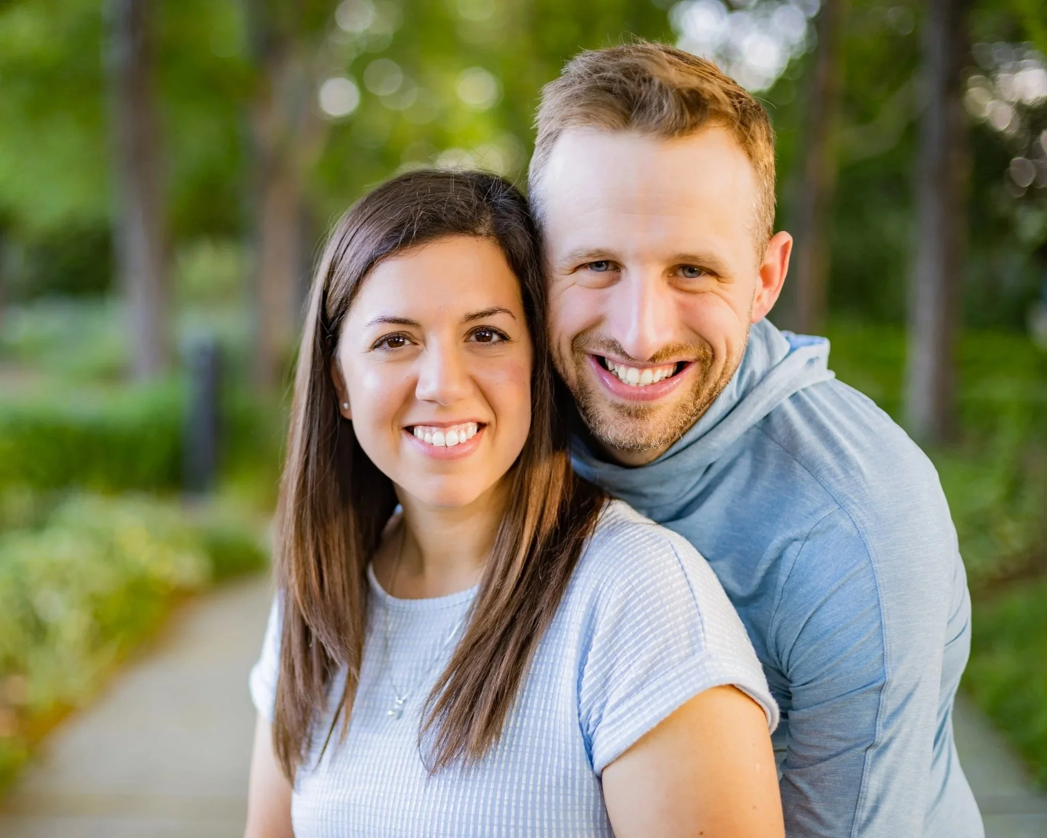 A smiling couple, a woman with long brown hair and a man with short blonde hair, standing outdoors with a blurred green background of trees, close together, face to face, happily smiling.