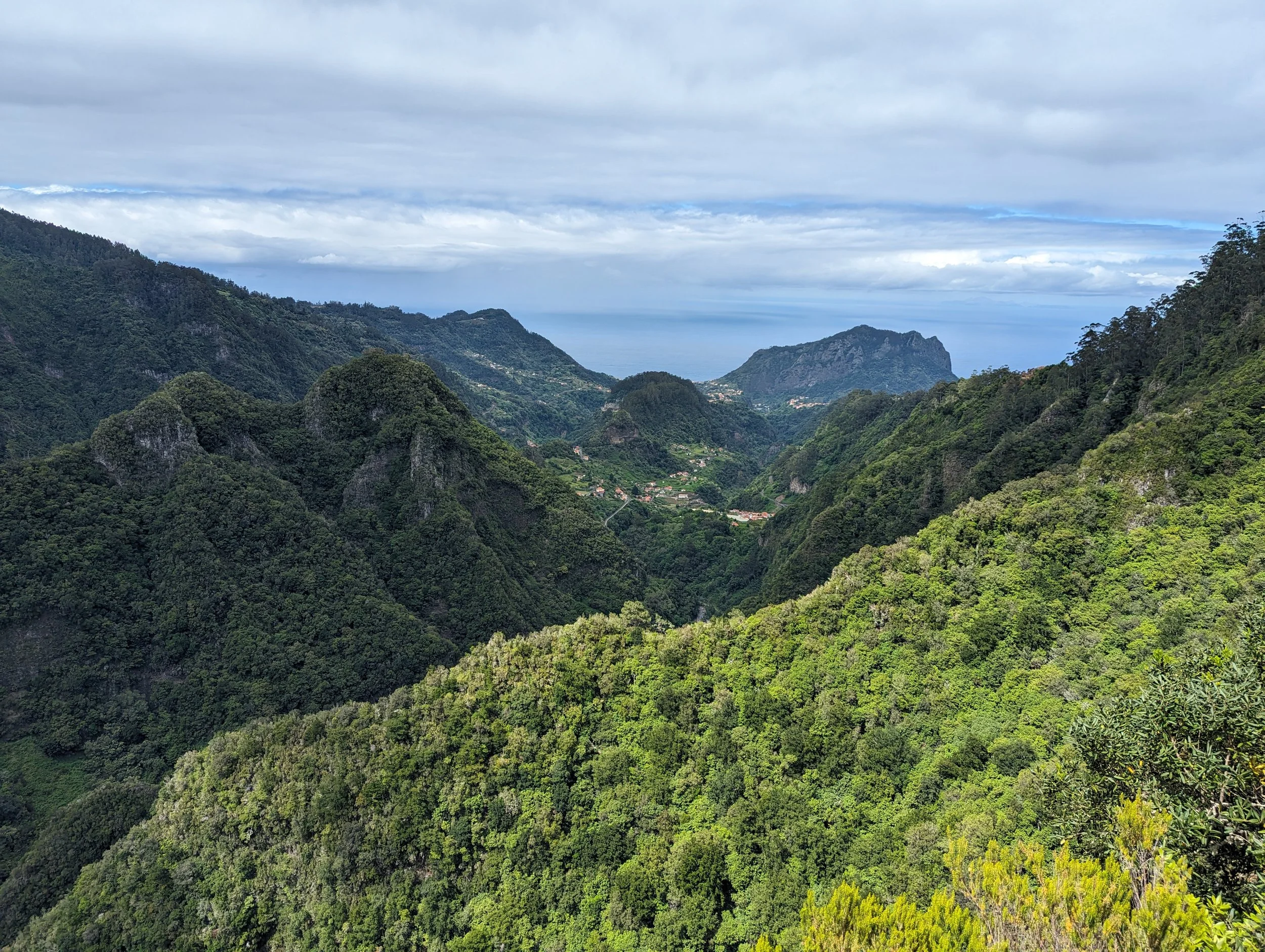 Scenic view of lush green mountains with scattered trees and small clusters of houses, under a cloudy sky over the ocean in the distance.