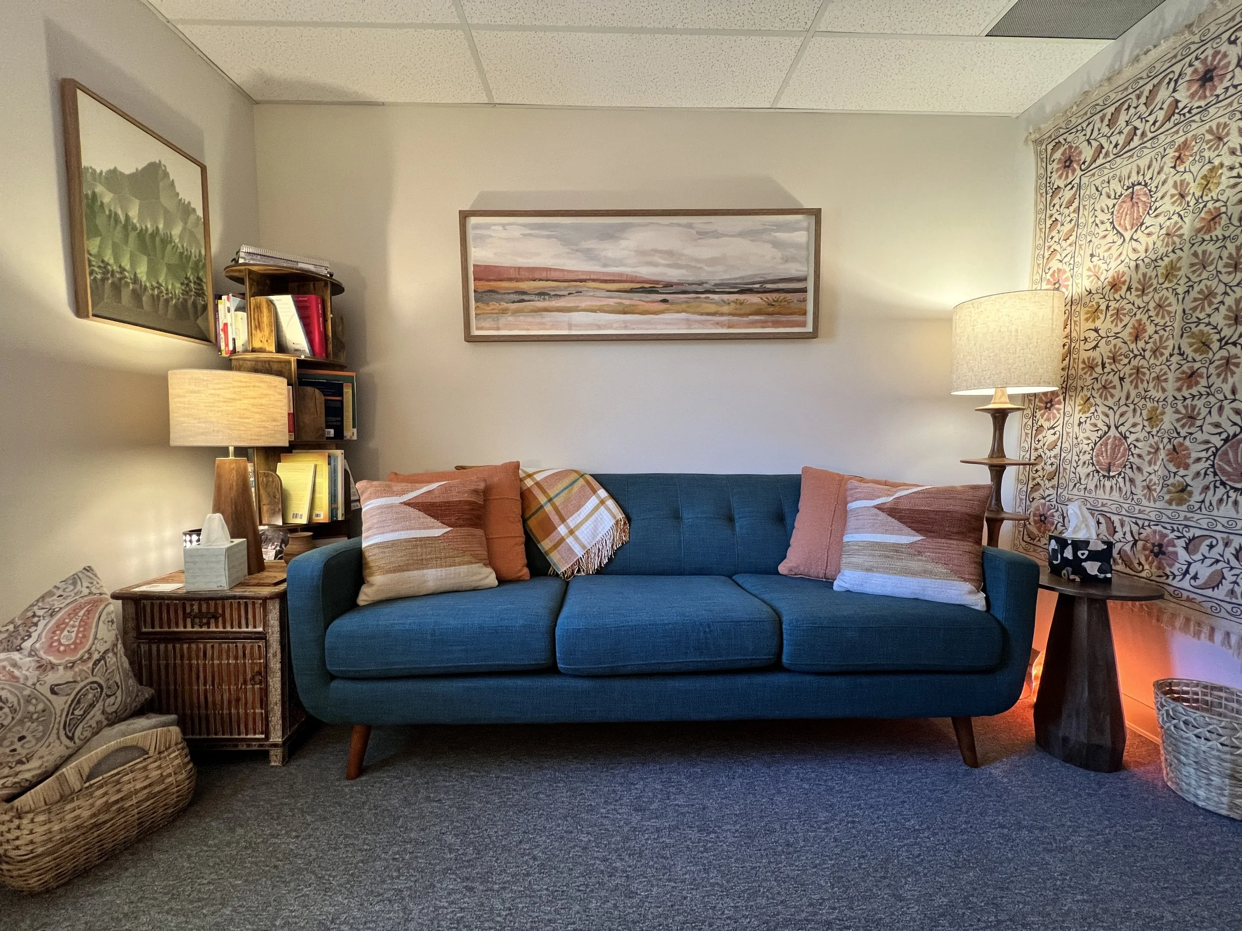 Living room with a teal sofa, decorated with square pillows and a blanket, flanked by two wooden side tables with lamps, a woven basket under each table, a patterned textile on the wall behind, and framed artworks on the walls.