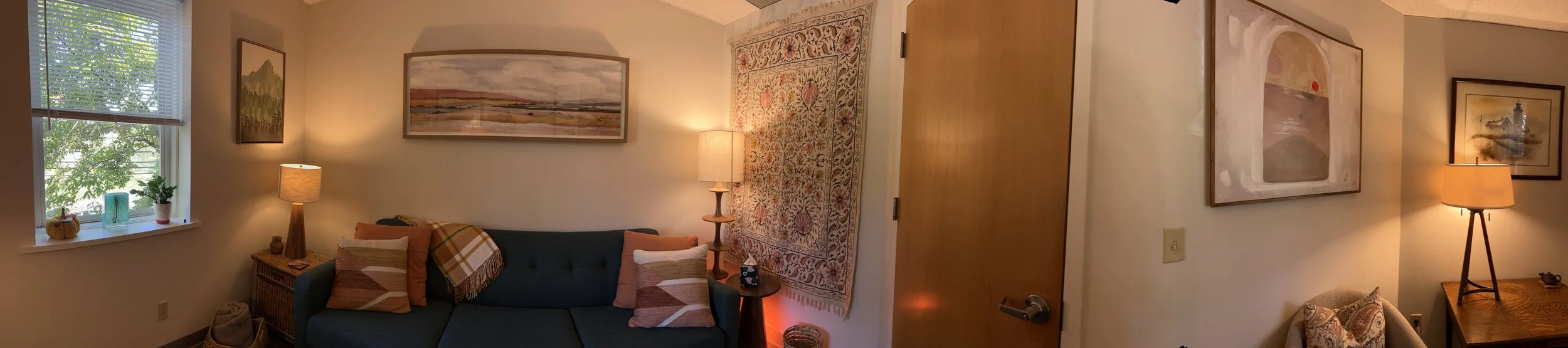 A cozy living room with a window on the left, decorated with potted plants and a small pumpkin. In the center, there's a blue sofa with plaid pillows and a blanket. Two lamps with warm lighting flank the sofa, and framed landscape artwork hangs on the wall above. To the right, there's a wooden door, a large decorative tapestry hanging on the wall, and another side table with a lamp and a framed picture.