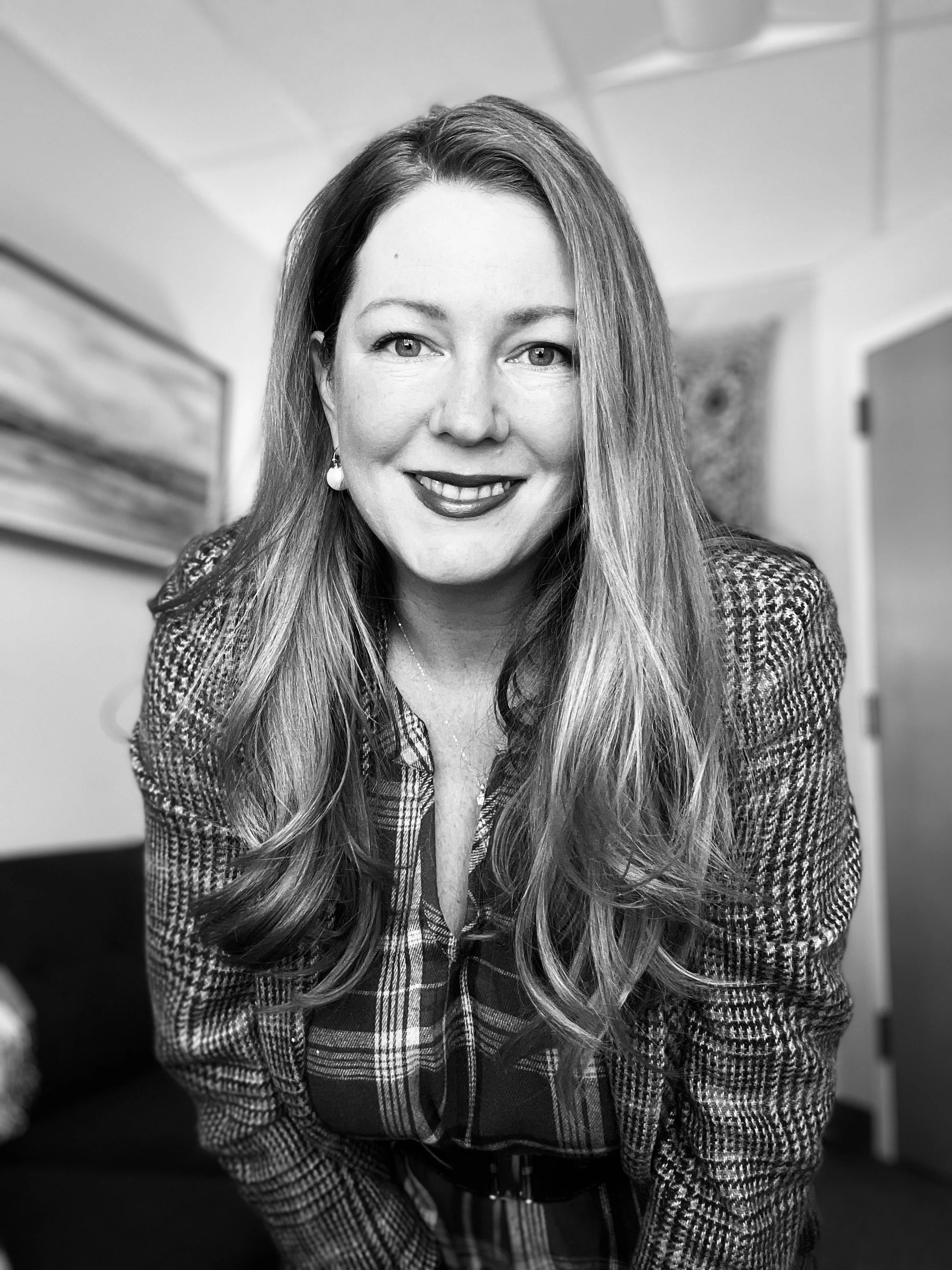 Black and white photograph of Anna Tivade LCSW, smiling with long hair, wearing a plaid shirt and earrings, in an office setting.