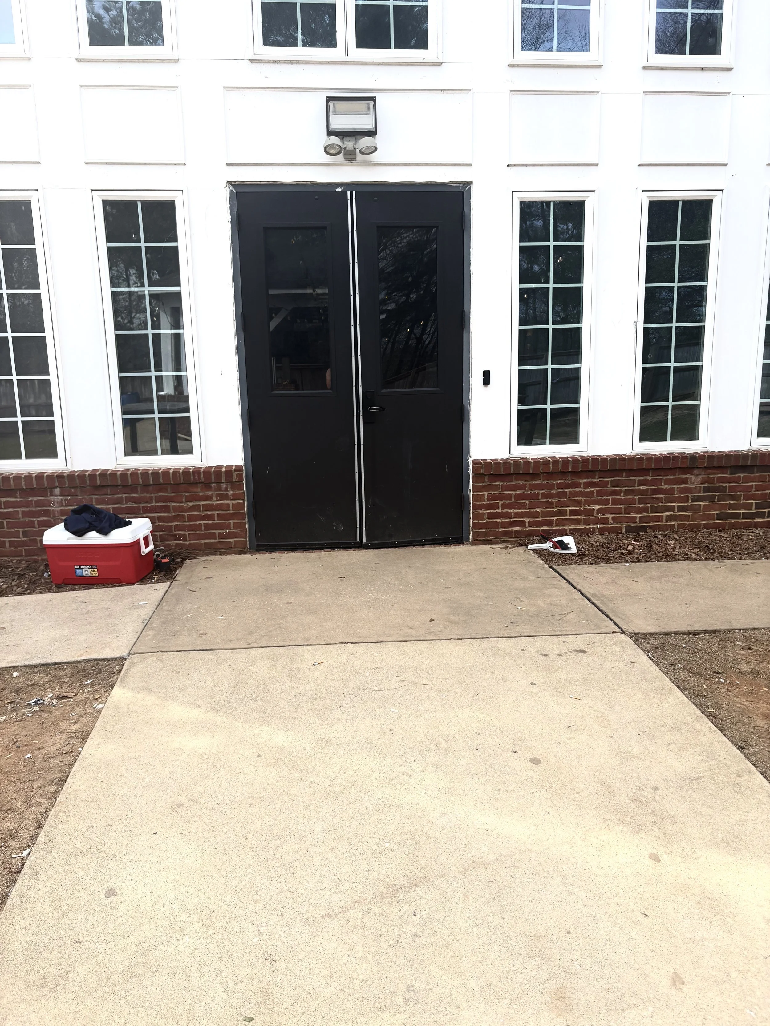 Front entrance of a building with black double doors, large windows, brick foundation, sidewalk, a cooler on the ground with clothes on top, and a tool on the ground to the right of the doors.