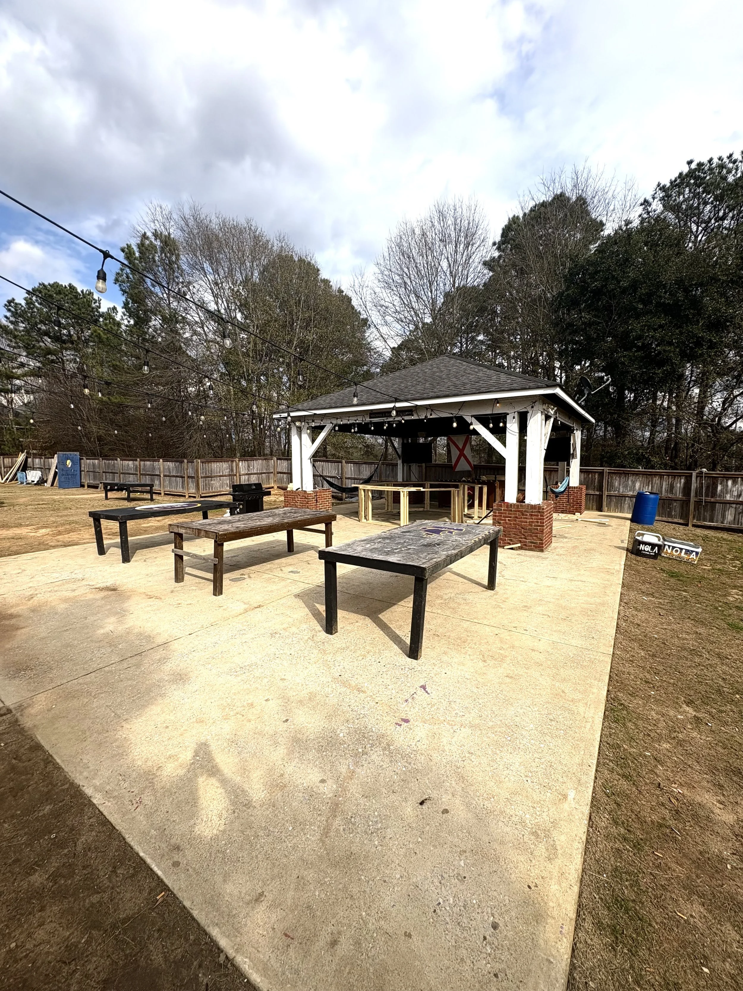 Outdoor patio with picnic tables, string lights, and a pavilion with a dartboard and an outdoor grill, surrounded by a wooden fence and trees.