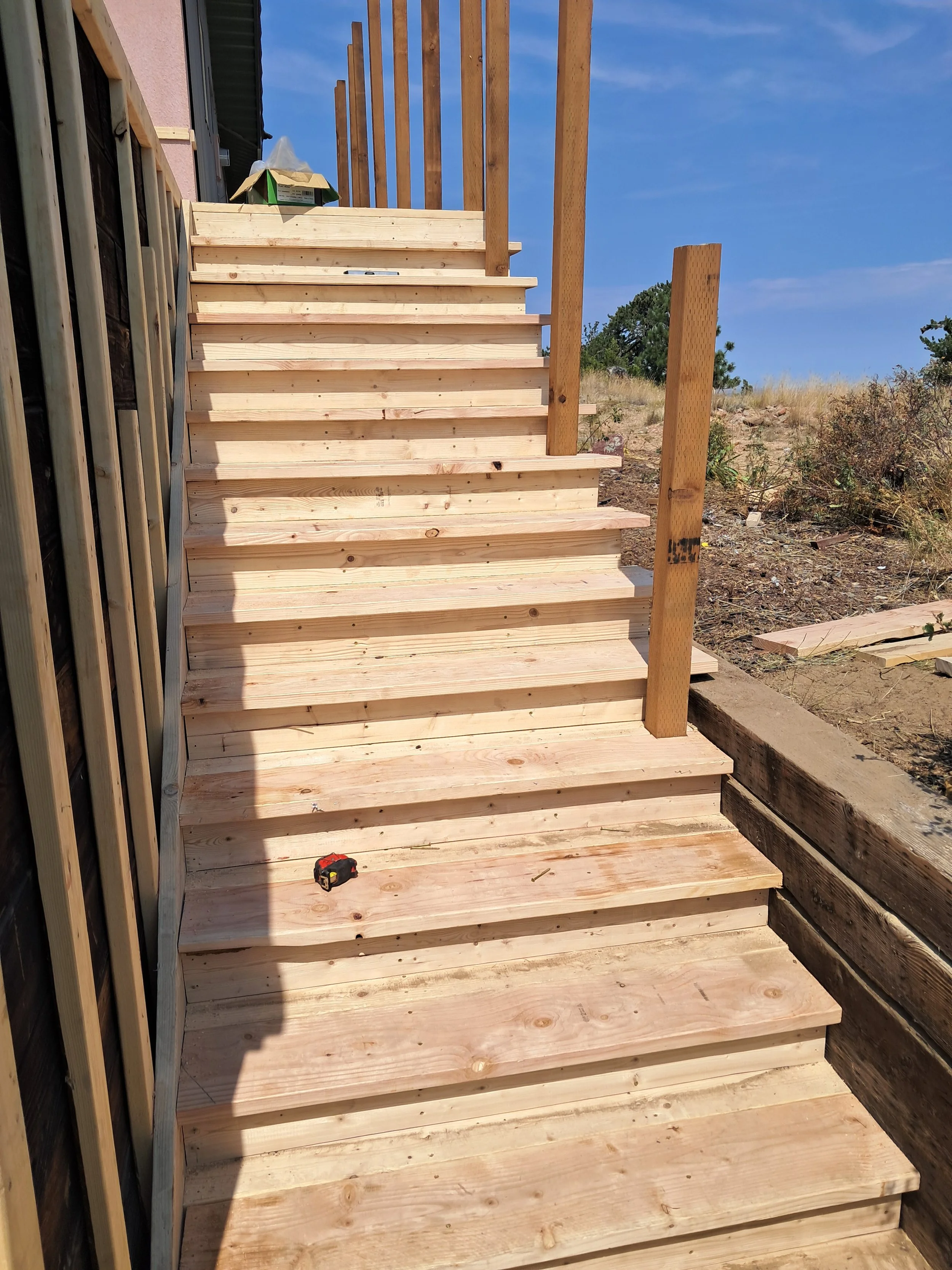 Wooden outdoor staircase under construction with new stairs and partial railing, on a sunny day, with a blue sky and some trees and bushes in the background.
