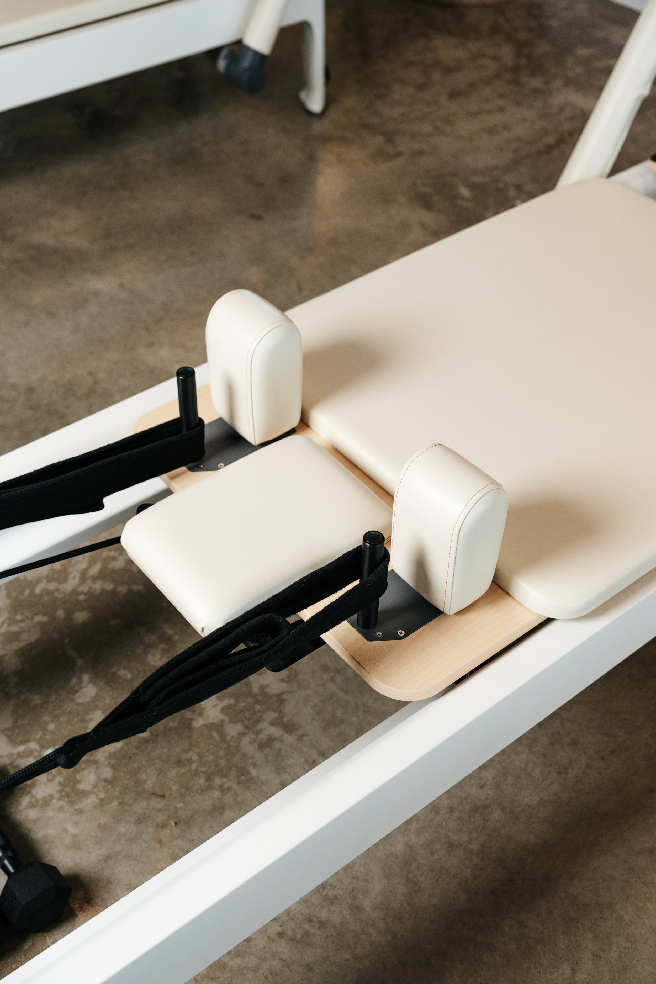 Close-up of a beige Reformer Pilates machine with padded supports and straps, placed on a concrete floor.