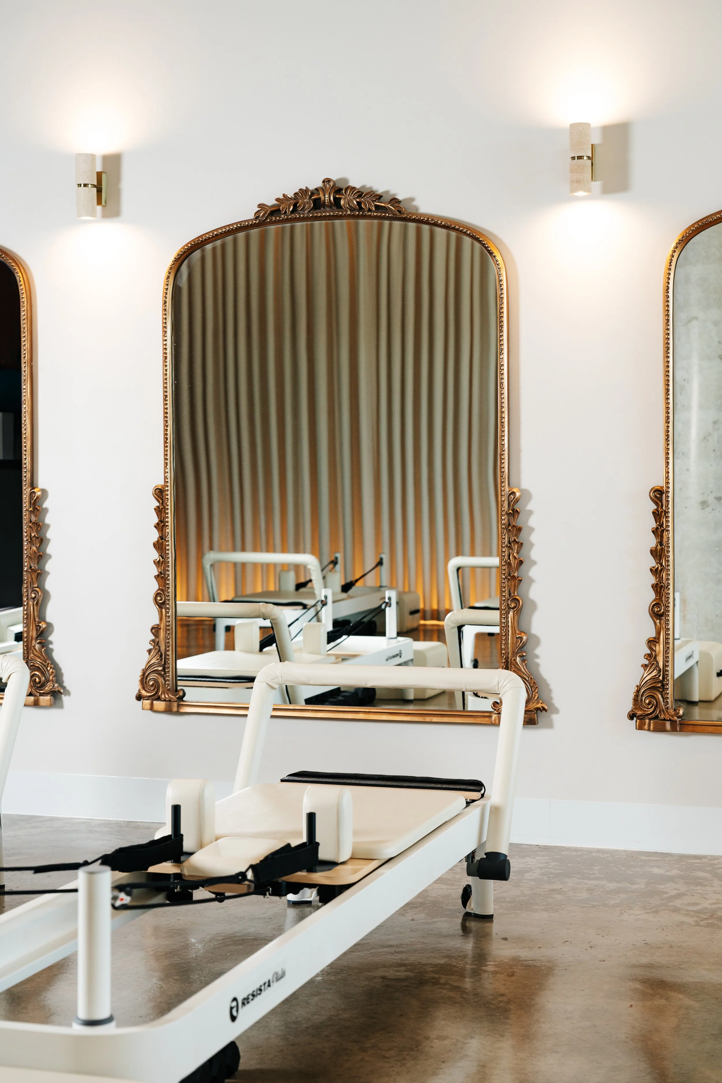 Reformer Pilates room with three ornate mirrors on the wall, beige curtains reflected in the mirrors, and beige Reformers with white frames and black straps in the foreground.