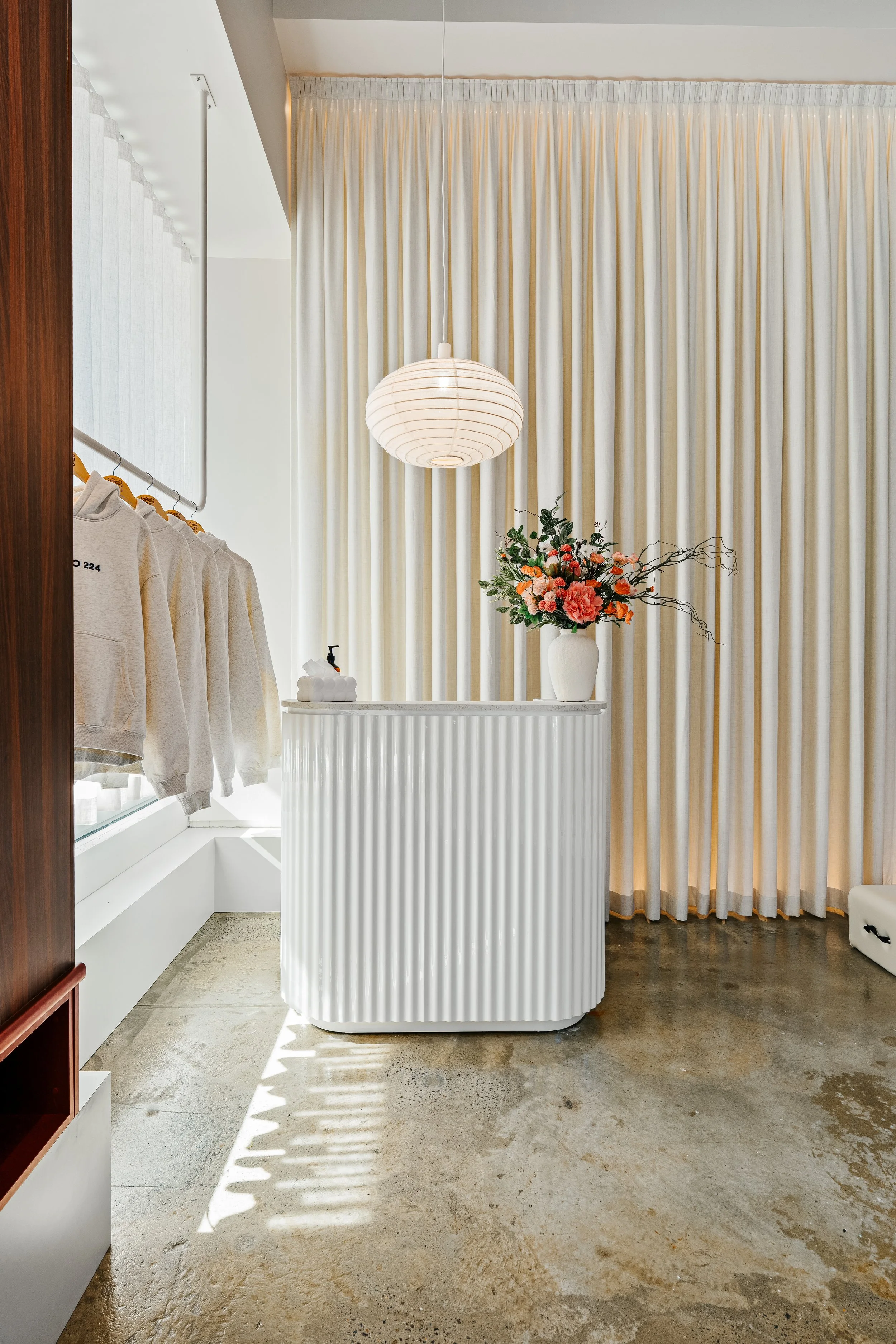 Interior of a room with beige curtains, a white reception counter with a vase of pink and orange flowers, and a white hanging paper lantern light fixture.