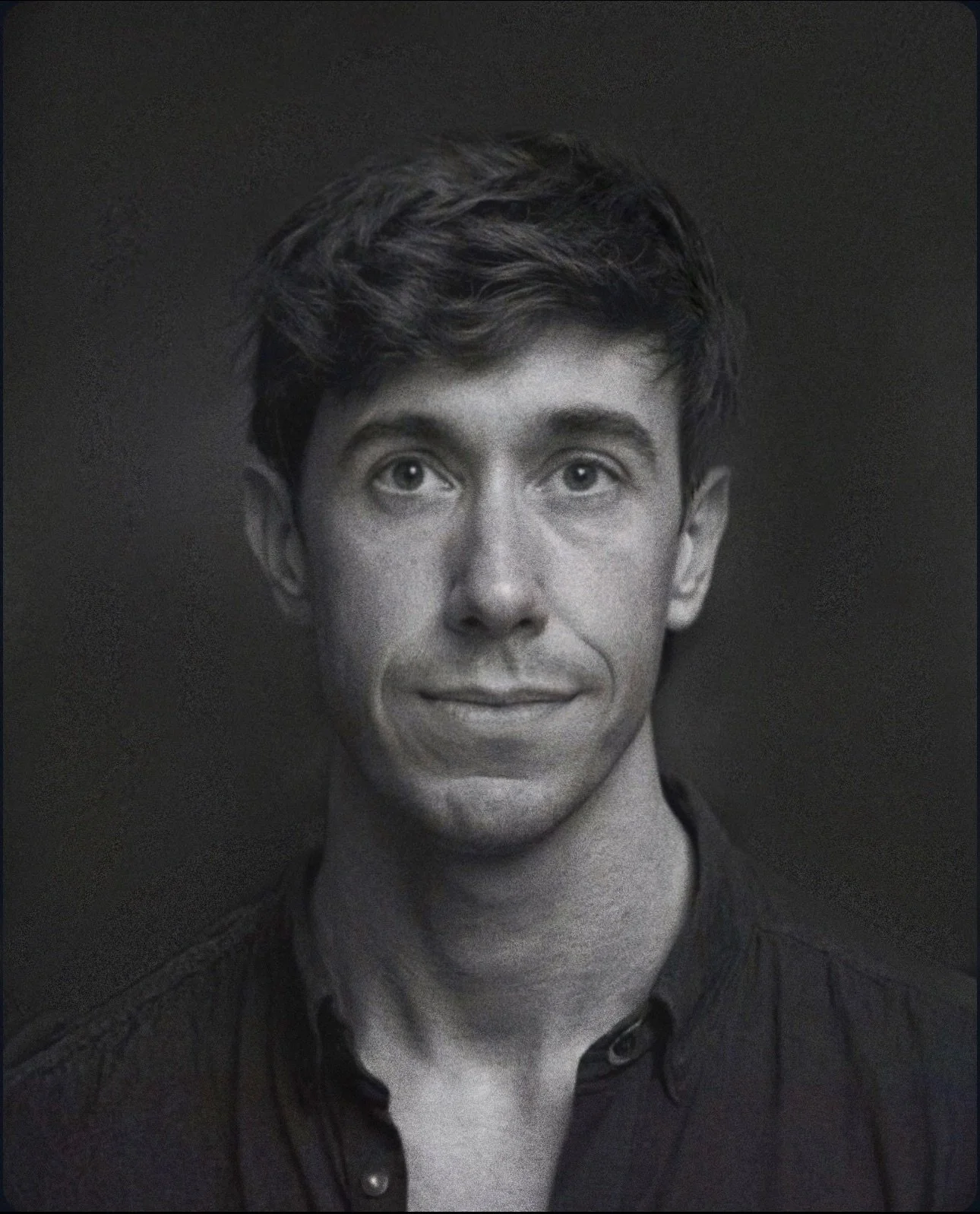 Black and white portrait of a young man with wavy hair, wearing a dark button-up shirt, looking directly at the camera with a slight smile.