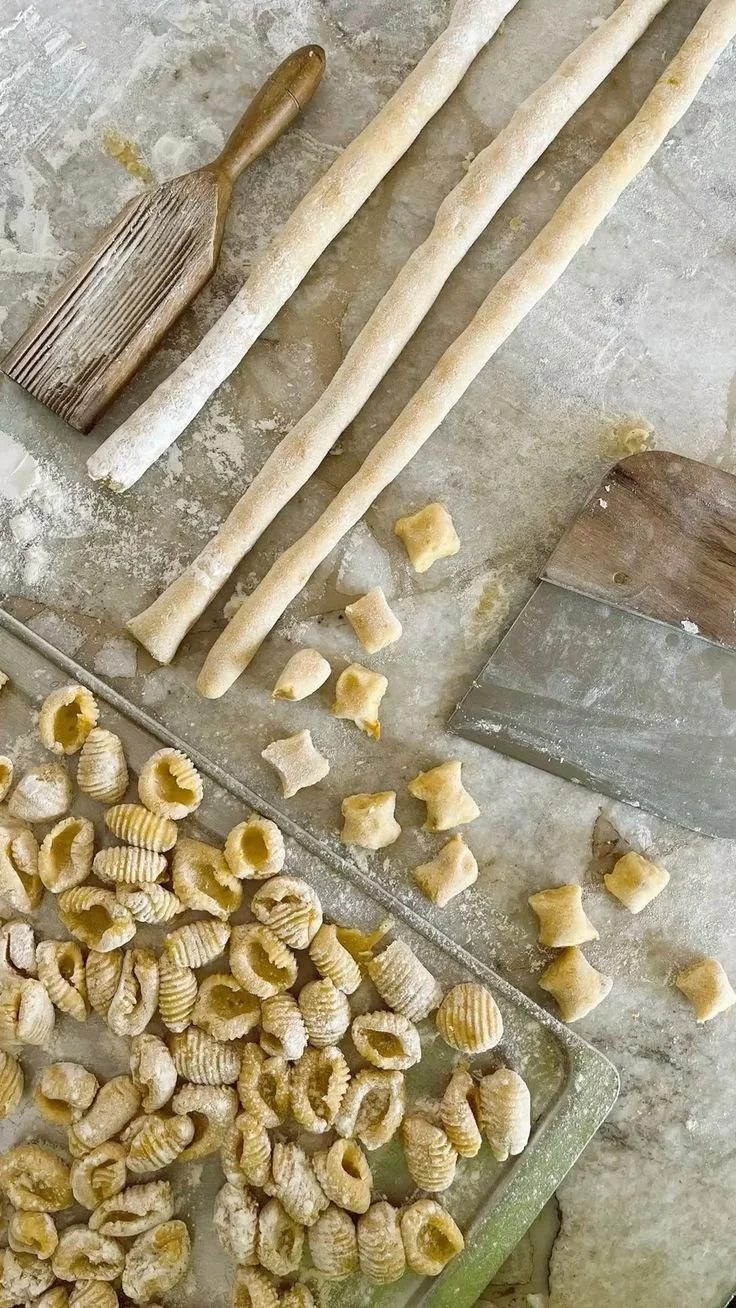 Uncooked pasta in various shapes and sizes, including long twisted strands, small star-shaped pieces, ridged shell pasta, and large shell pasta on a floured surface with a dough scraper, a pastry brush, and a grater nearby.