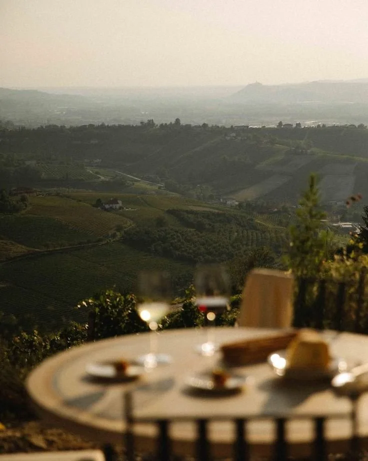A table with two wine glasses, a small dish, and a bowl of cheese set outdoors with a panoramic view of rolling green hills and vineyards at sunset.