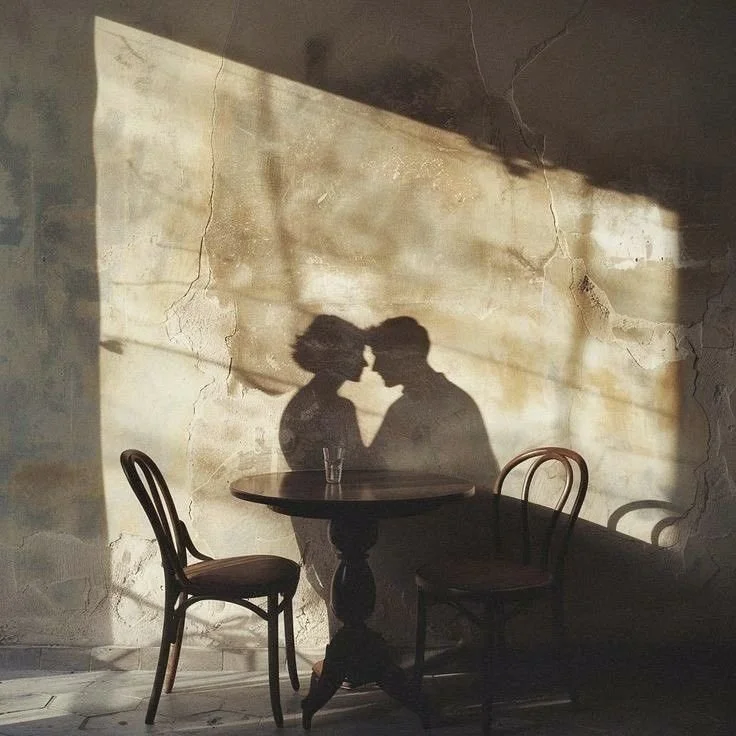 Shadow of a couple leaning in for a kiss on a wall with cracks, behind a wooden table with two chairs.