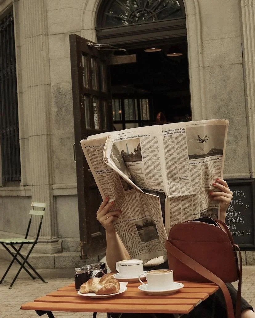 Person sitting at an outdoor cafe reading a newspaper, with a camera, coffee cups, croissants, a brown bag, and a blackboard menu on the table. The background shows a building entrance and a black open doorway.