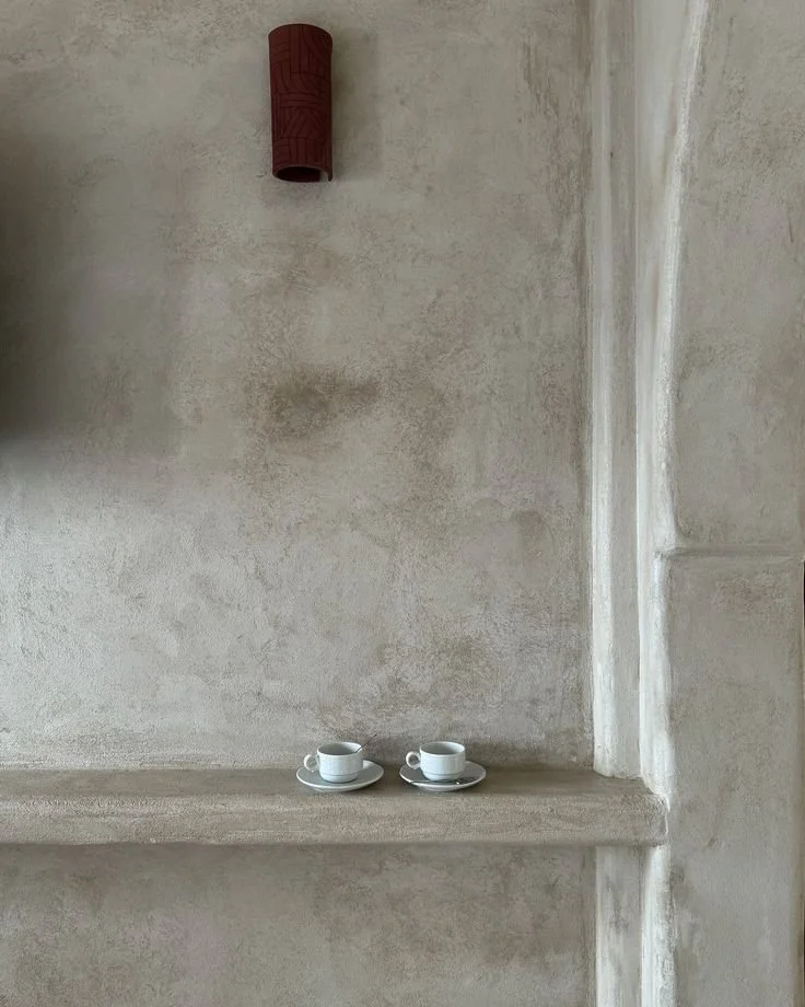 Two small white coffee cups with saucers placed on a light-colored shelf against a textured beige wall.