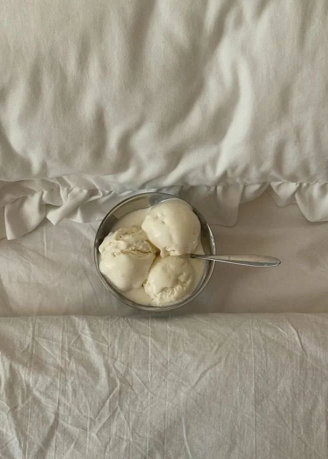A small metal bowl with three scoops of vanilla ice cream and a spoon on a white tablecloth, with a white pillow and bed linen in the background.